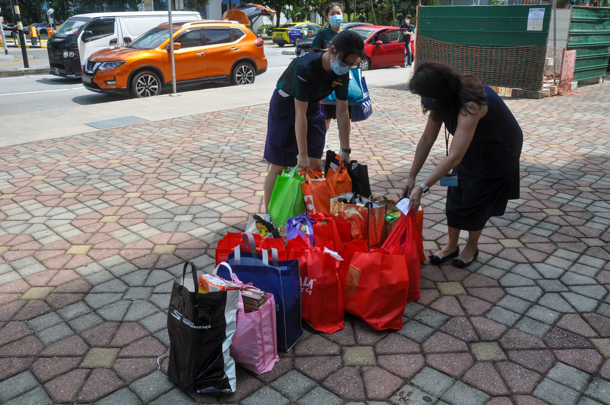 Two masked people sort many colorful tote bags on a paved sidewalk near a busy road with parked cars.