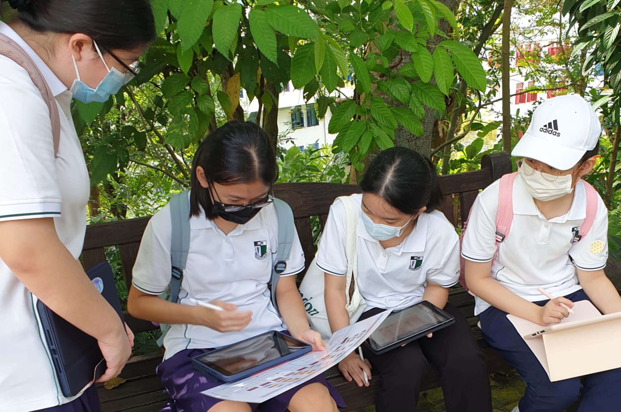 Four masked students in white uniforms sit on an outdoor bench, reviewing papers and tablets under green trees.