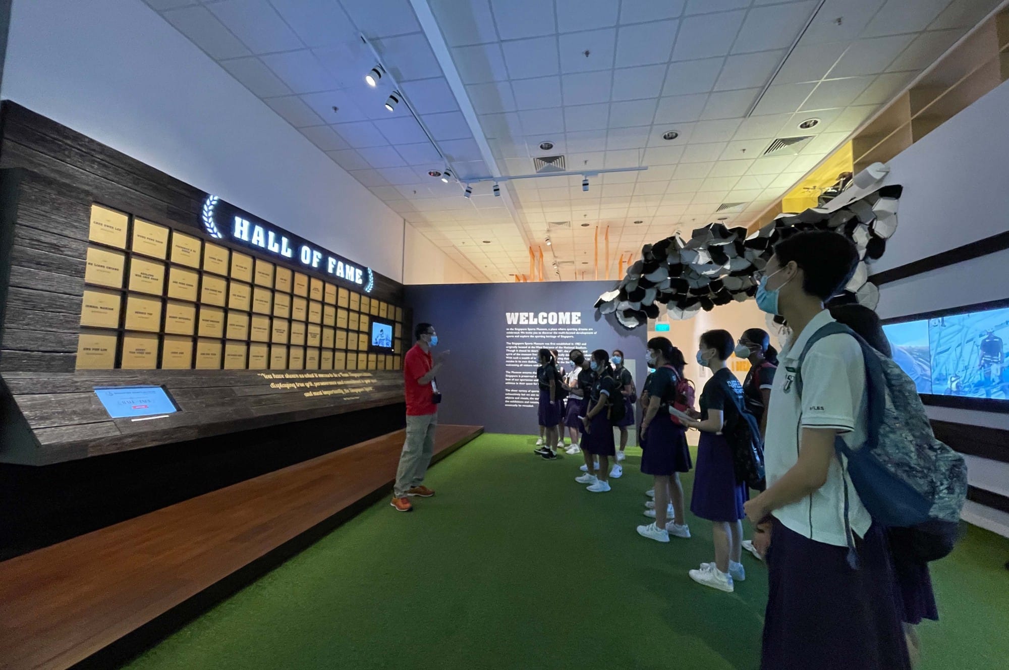 Students wearing masks tour a Hall of Fame exhibit with gold plaques on a dark wooden wall.