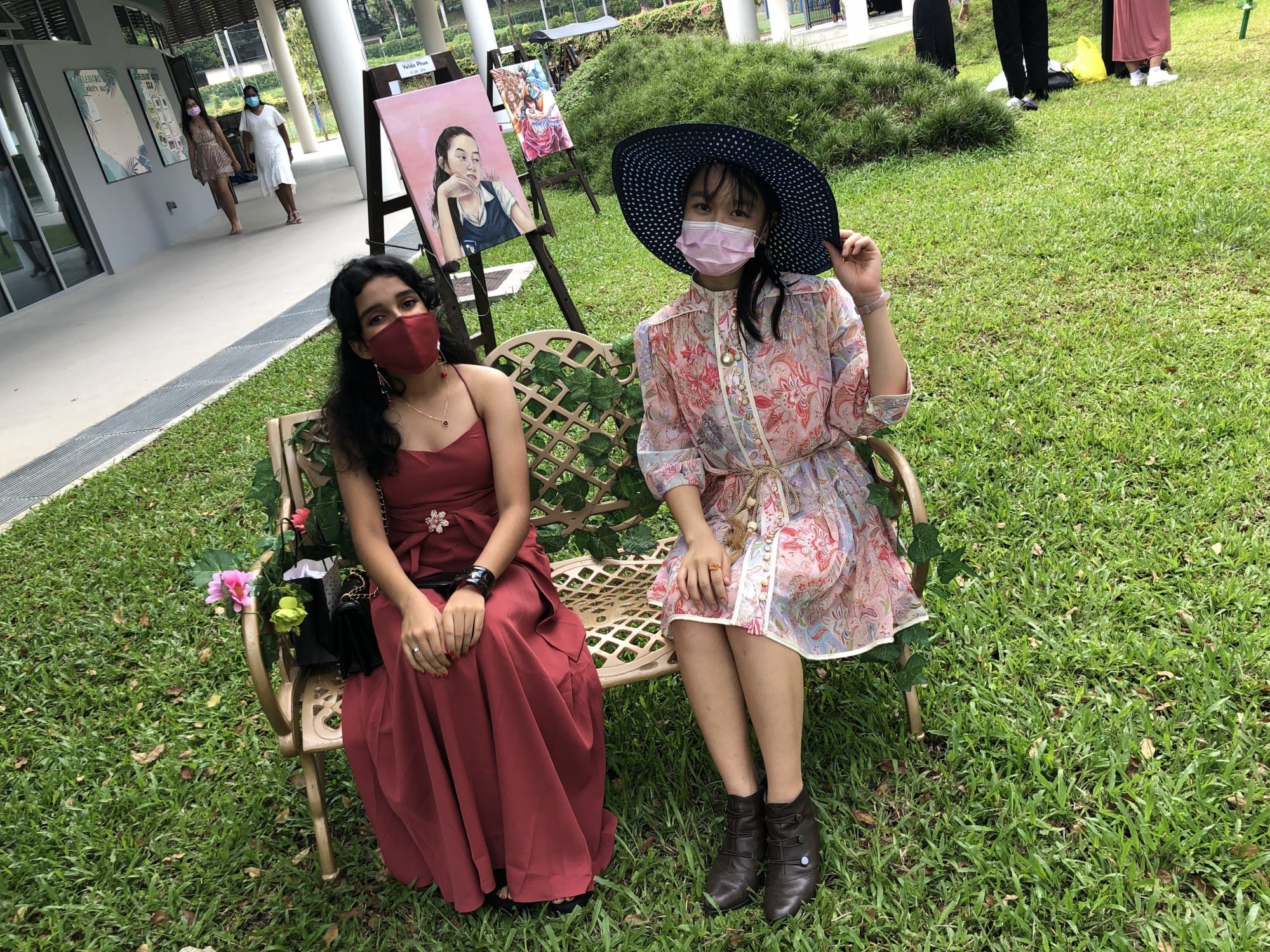 Two masked women sit on an outdoor bench near displayed paintings on grass, one in red dress, one in floral dress and wide hat.