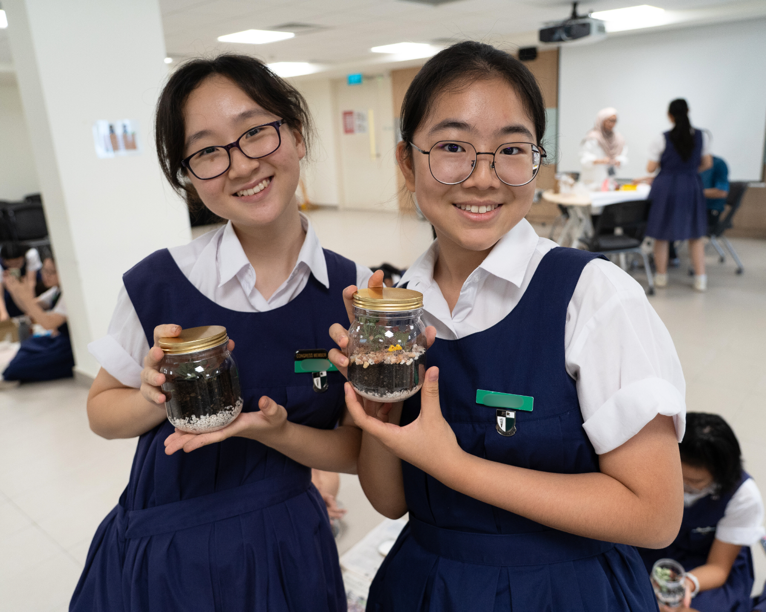 Two smiling schoolgirls in navy uniforms hold glass jars filled with layered soil and pebbles in a classroom.