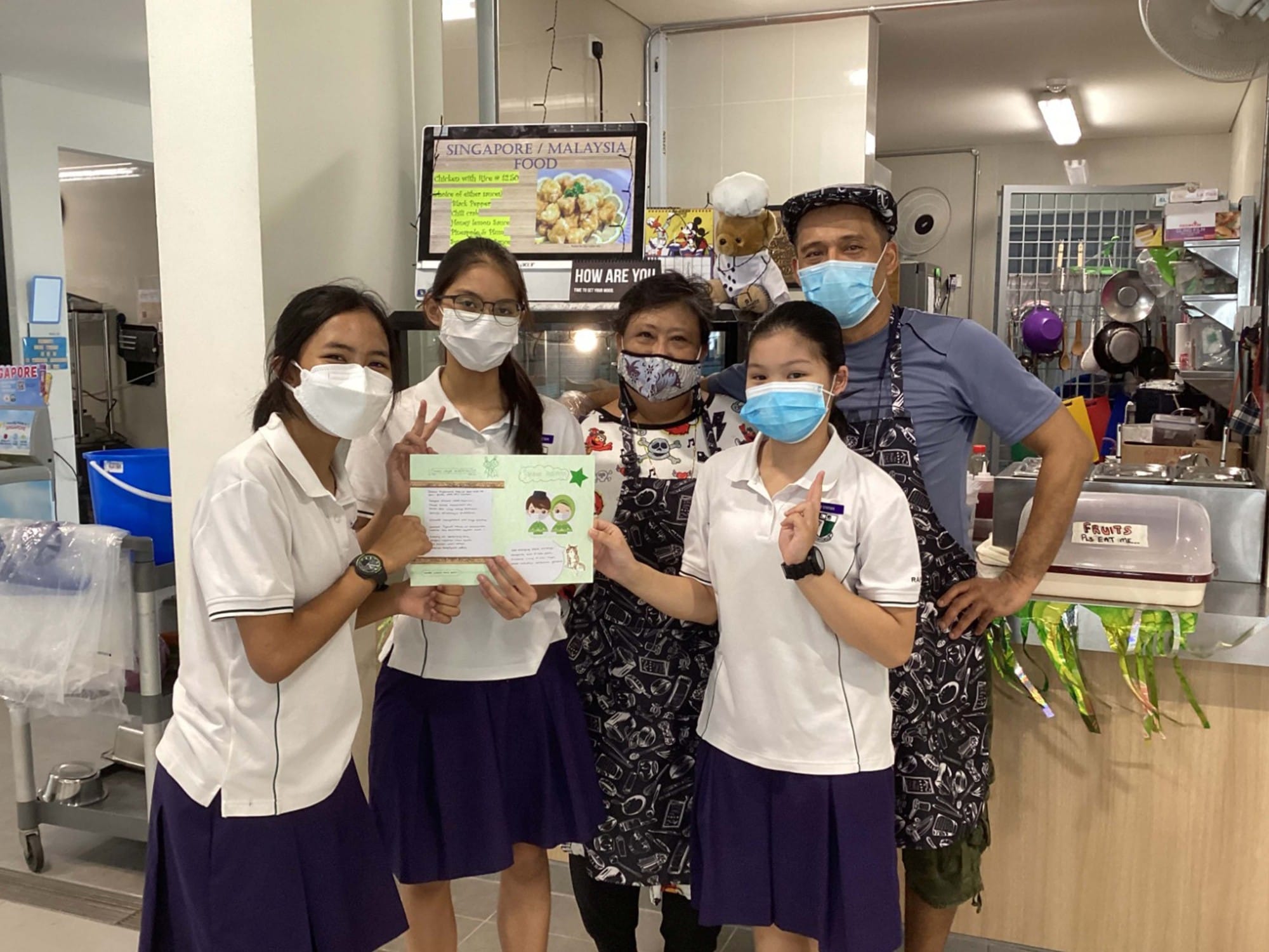 Five people wearing face masks pose together in a food stall, with students holding a handmade card.