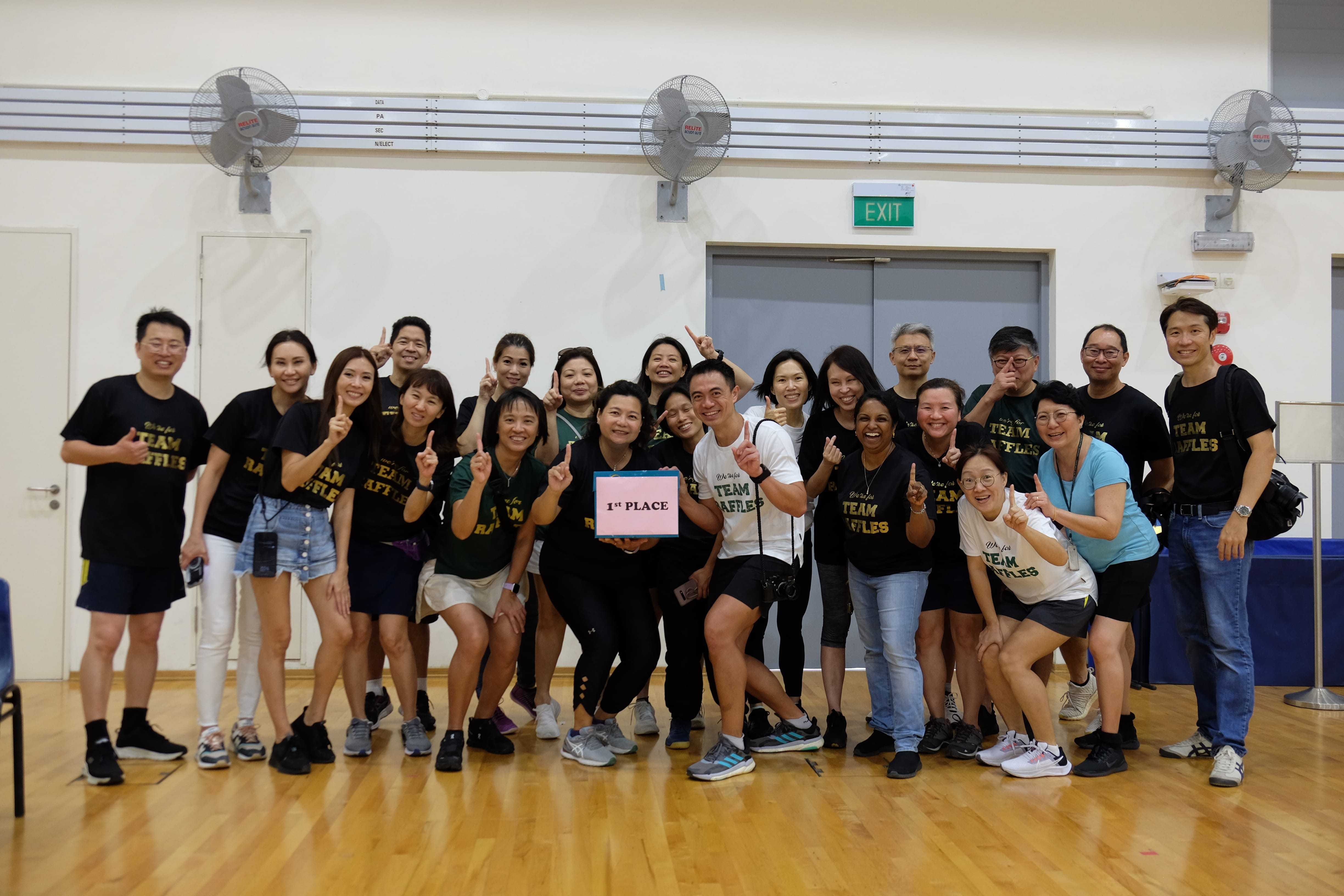 Group of people posing in a gym, many holding up one finger, one person holds a 1st place sign.