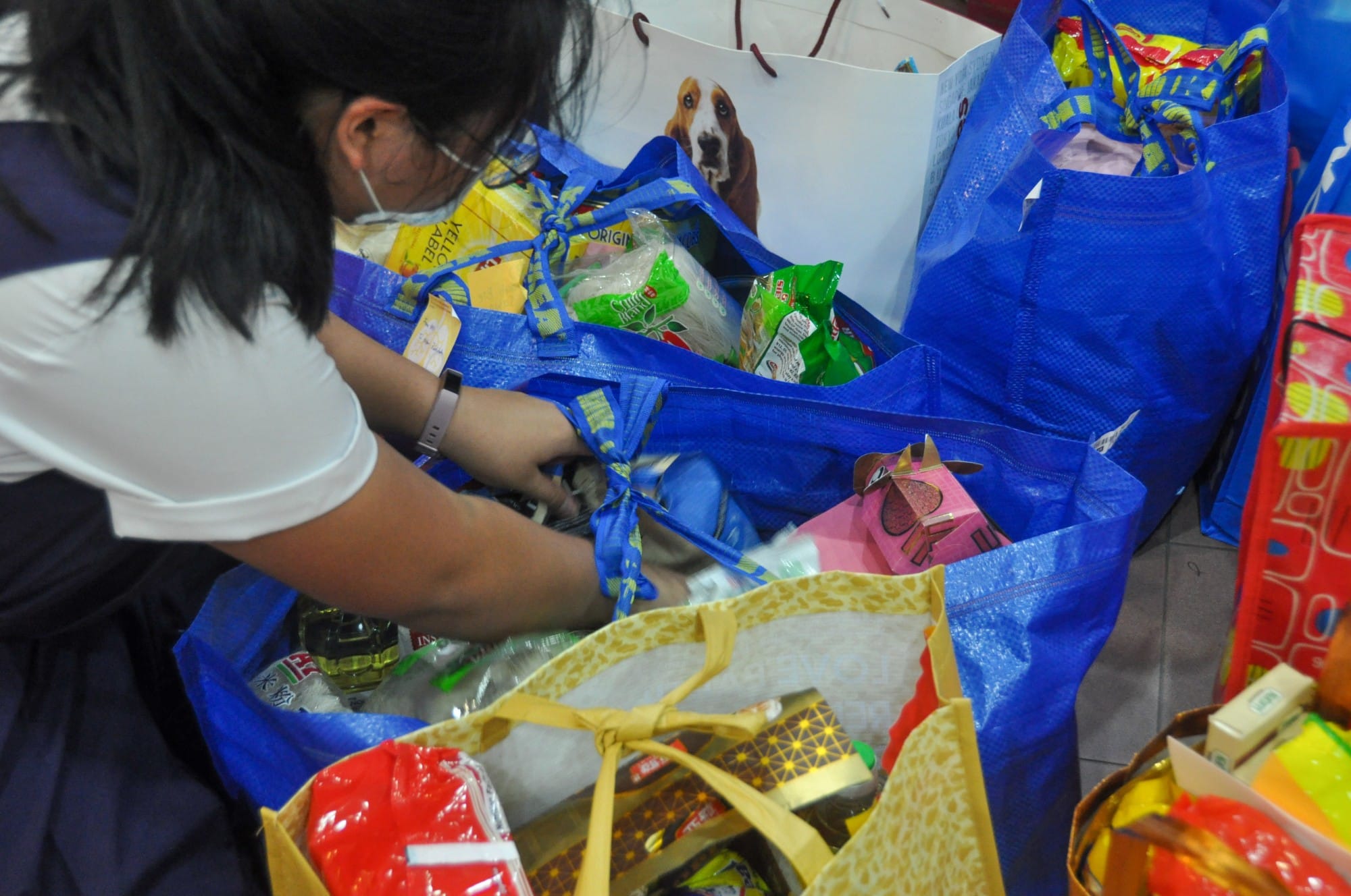 Person wearing a mask sorting through multiple large blue and yellow reusable bags filled with groceries and packaged goods.