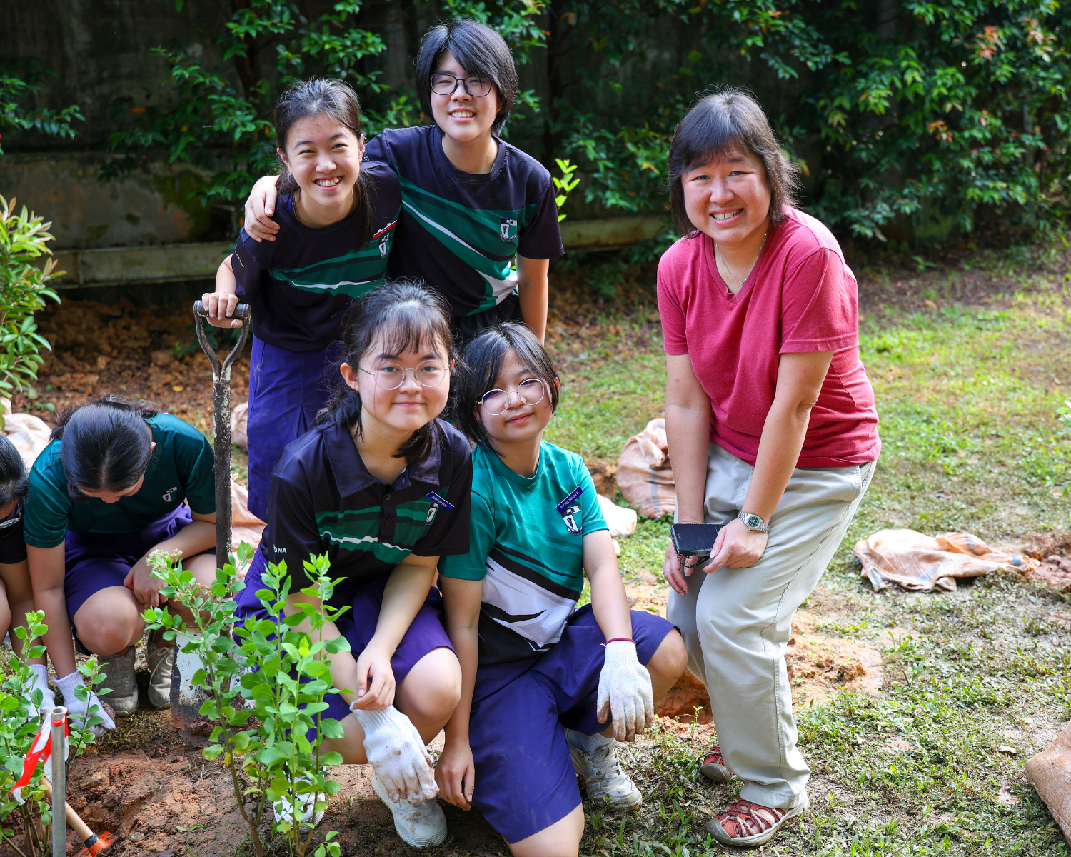 Five people smile while planting a sapling outdoors; one kneels with a shovel, another holds a phone.
