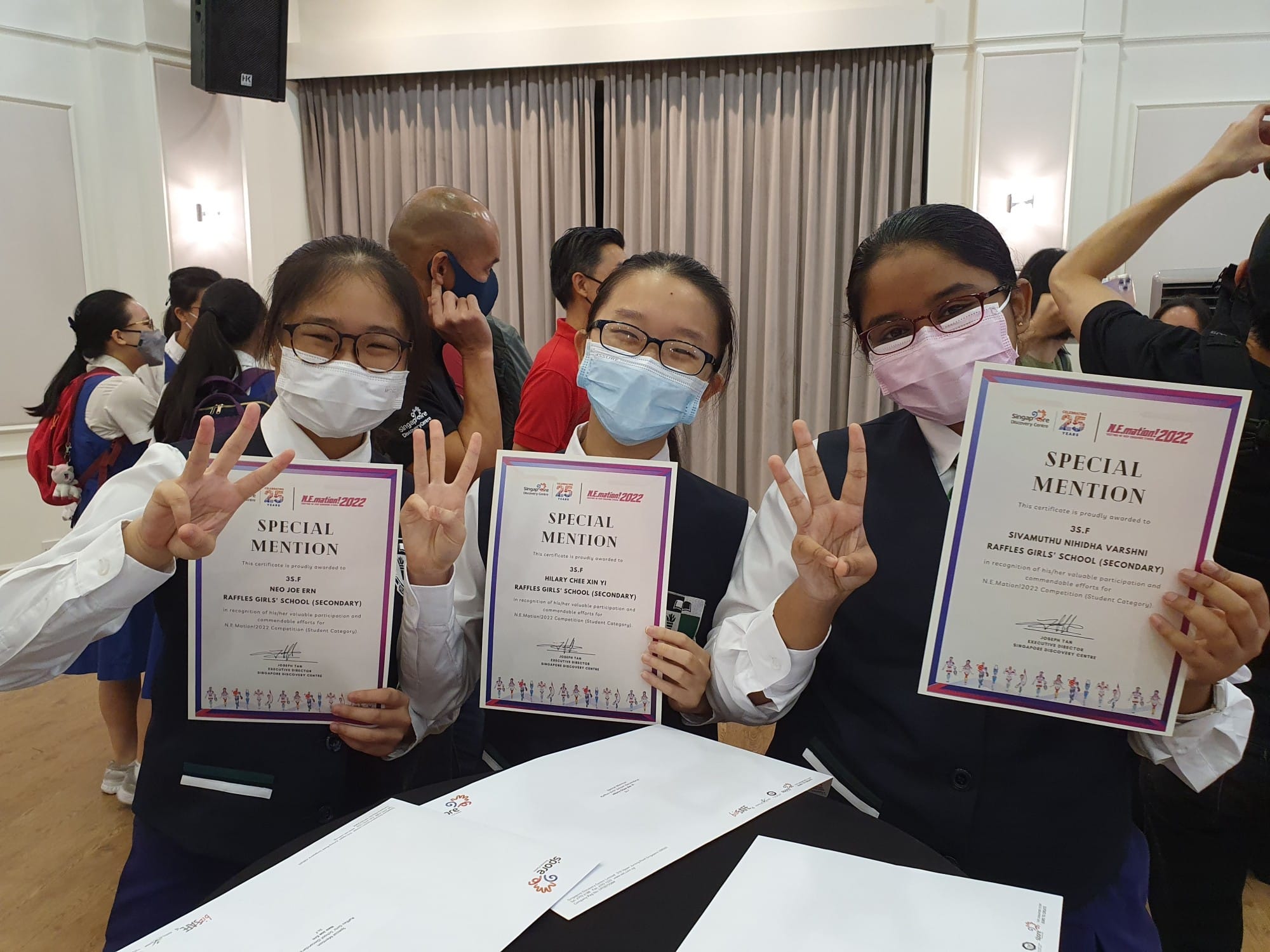 Three masked schoolgirls hold Special Mention certificates from N.E.motion 2022, flashing peace signs.