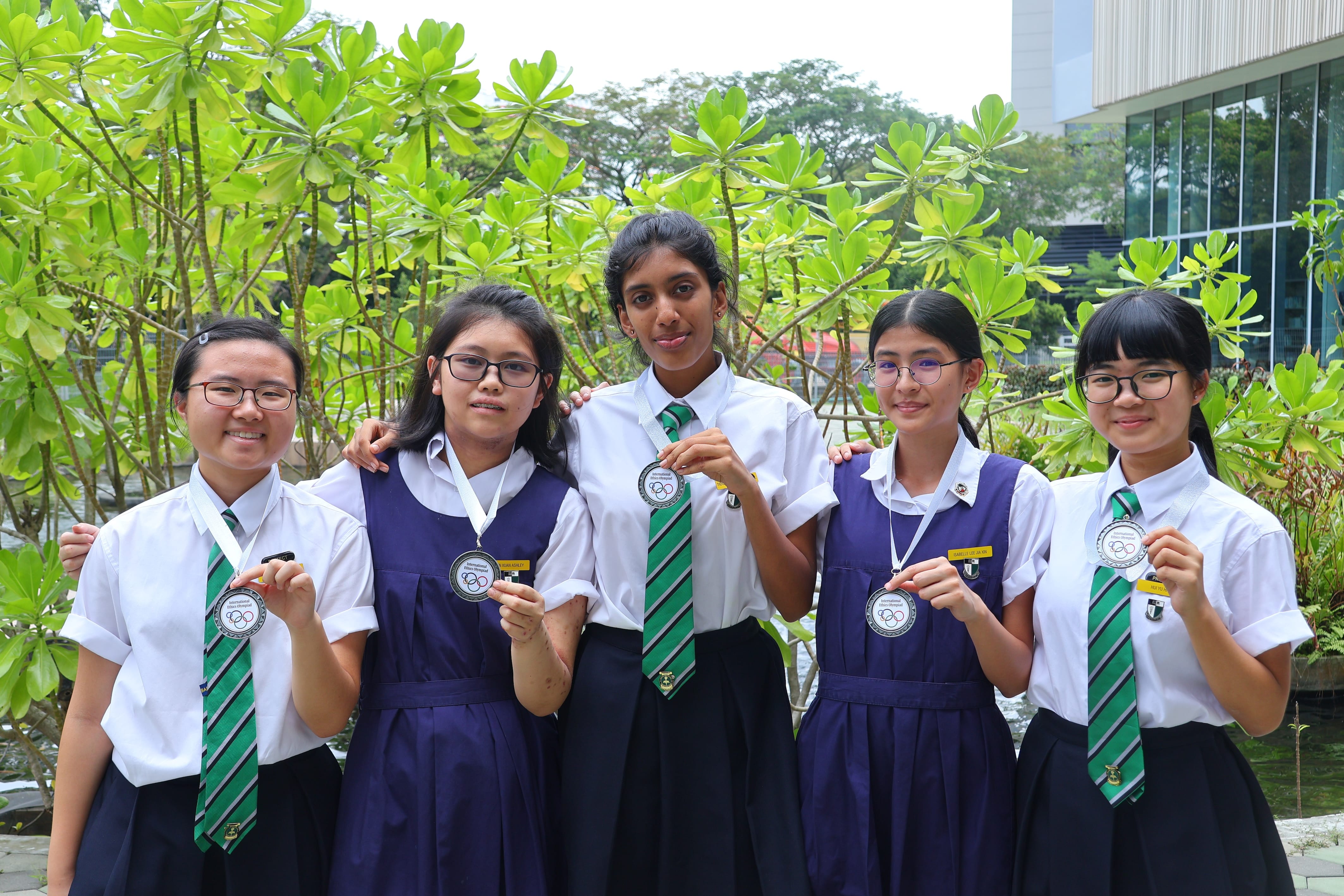 Five schoolgirls in uniforms with green ties smile and hold medals outdoors near green trees.