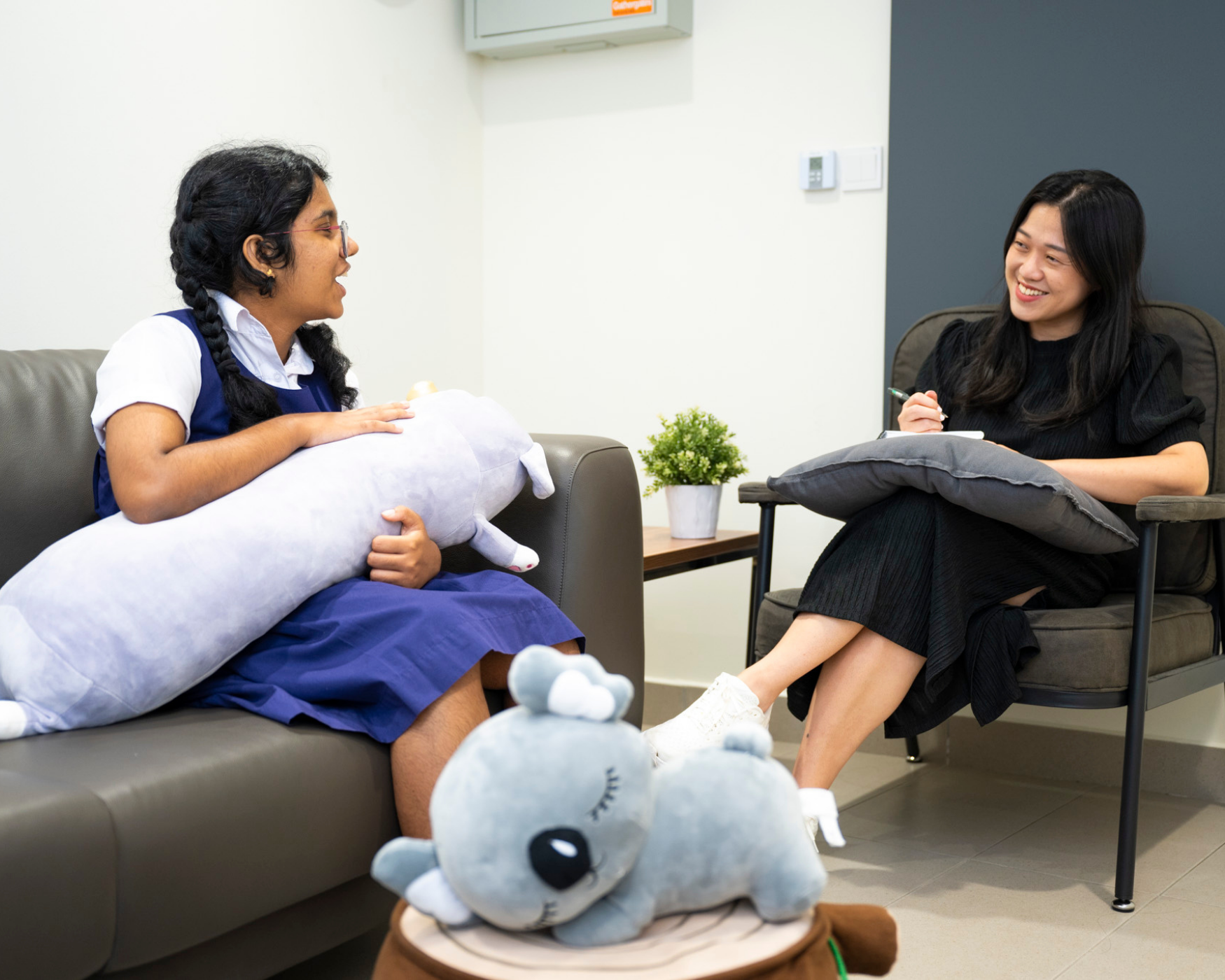 A young girl holding a plush toy talks to a smiling woman with a clipboard in a cozy counseling room.