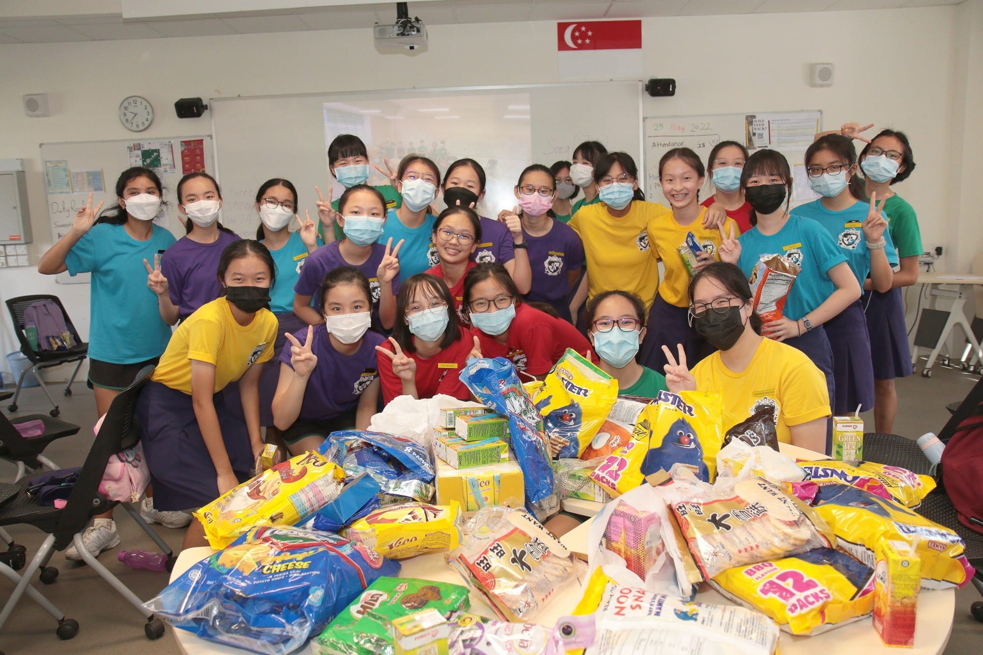 A large group of masked students in colorful uniforms pose with peace signs behind a table piled with donated food items.