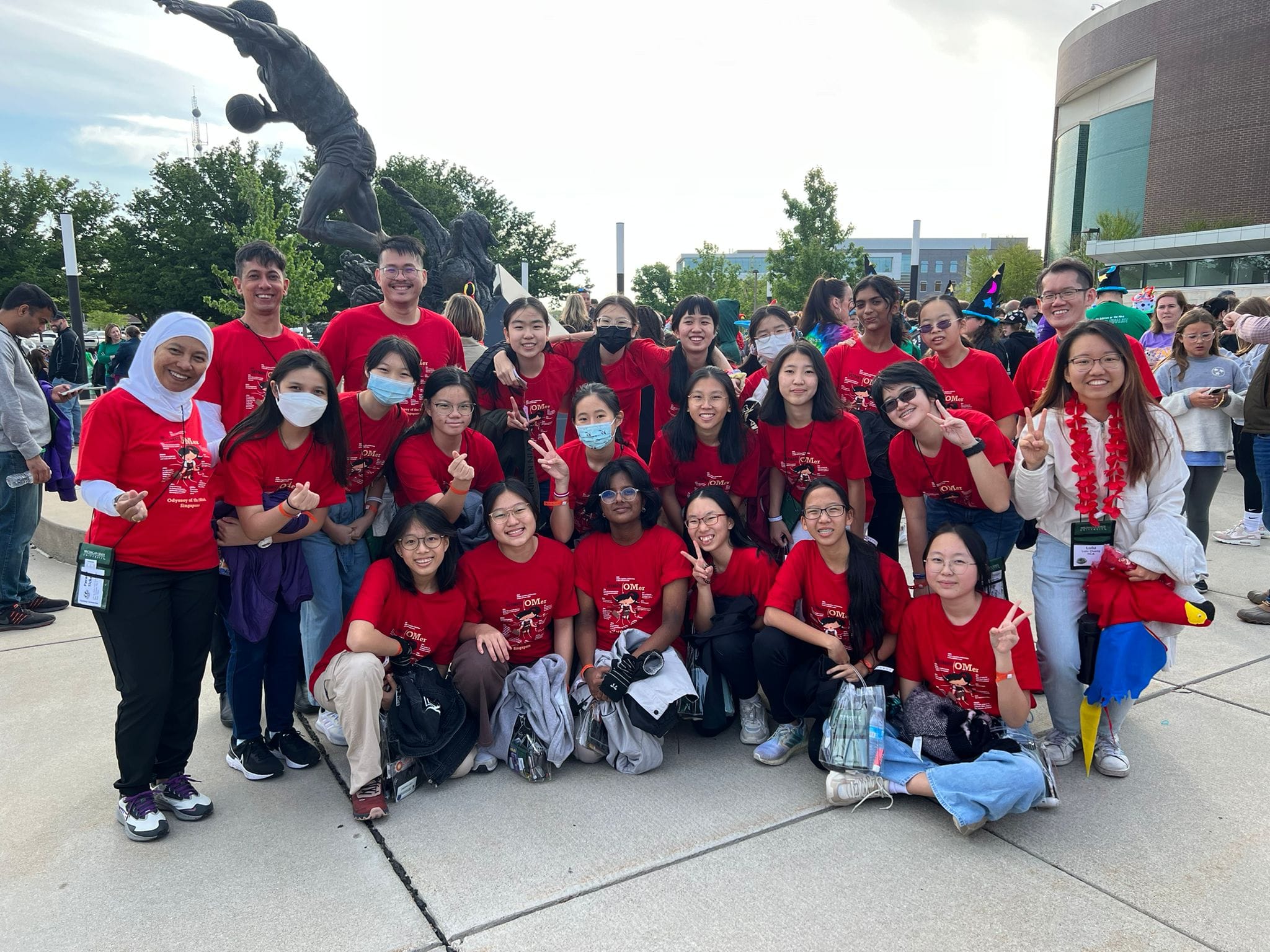 A large group of young people in matching red shirts pose outdoors near a bronze athlete statue.