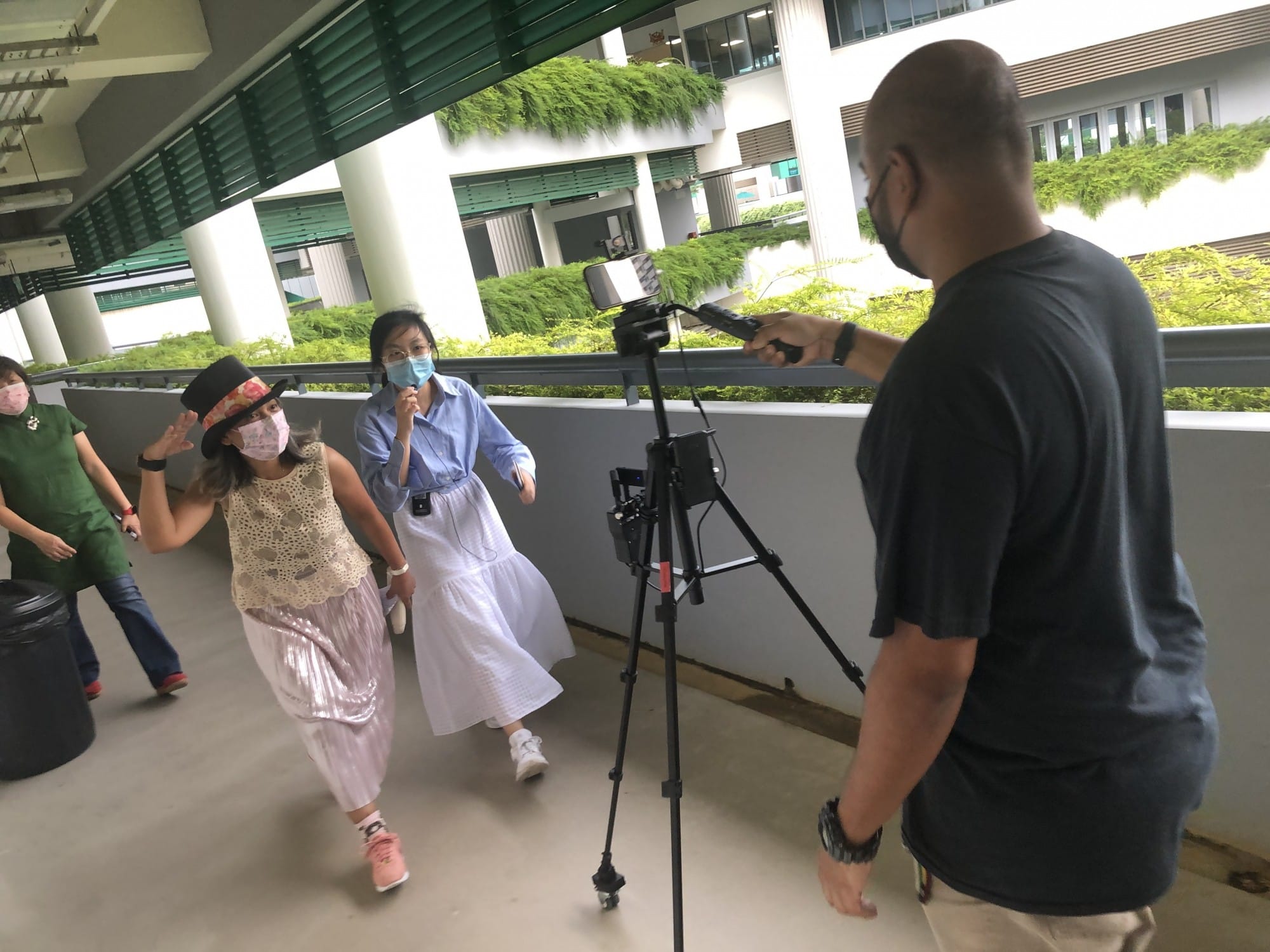 A man films three masked women walking toward a camera on a tripod on an outdoor corridor of a building.