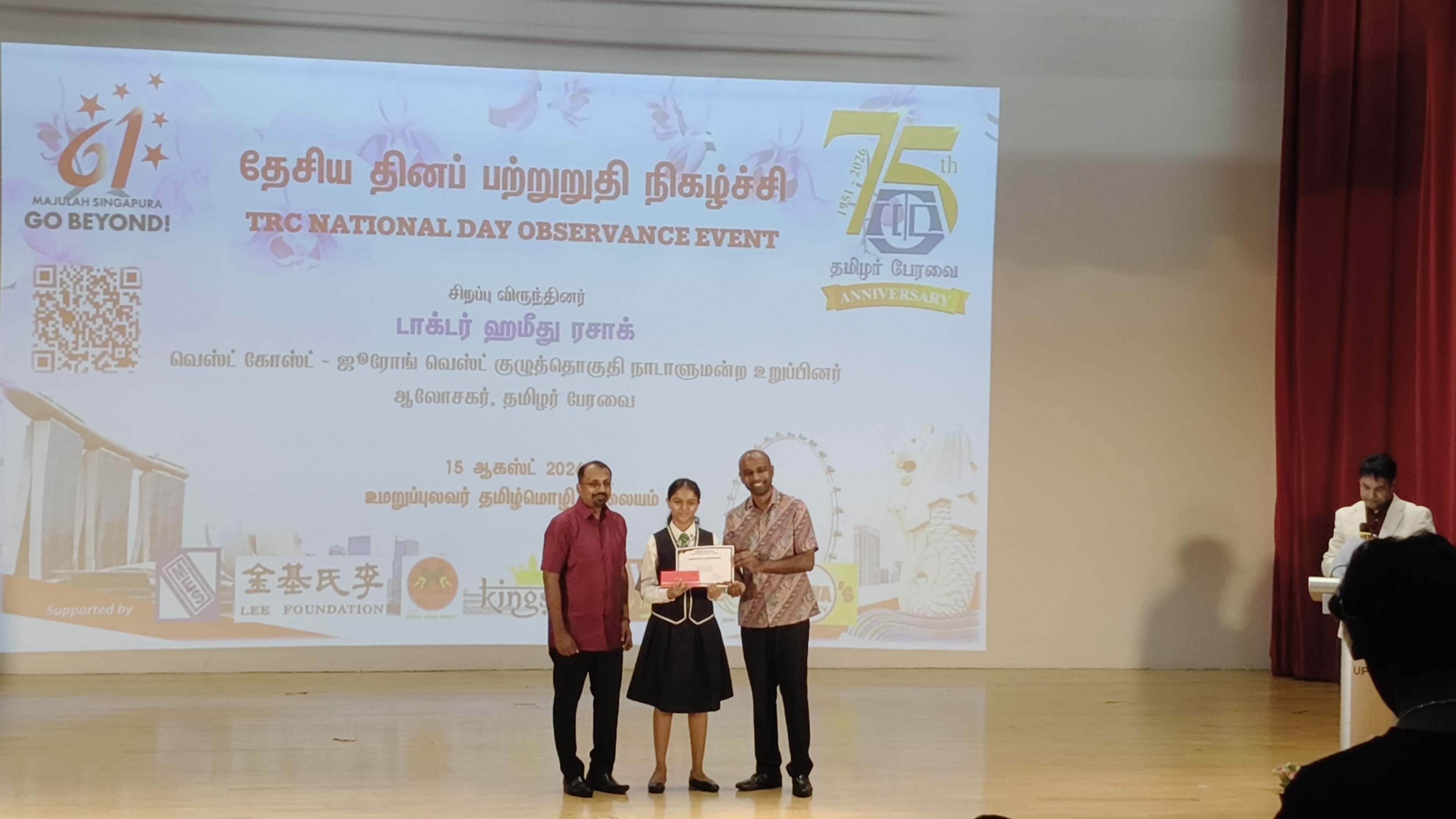 A female student standing on stage with a certificate presented to her with another accompanying adult beside her, against a backdrop of a projector screen titled "TRC National Day Observance Event".