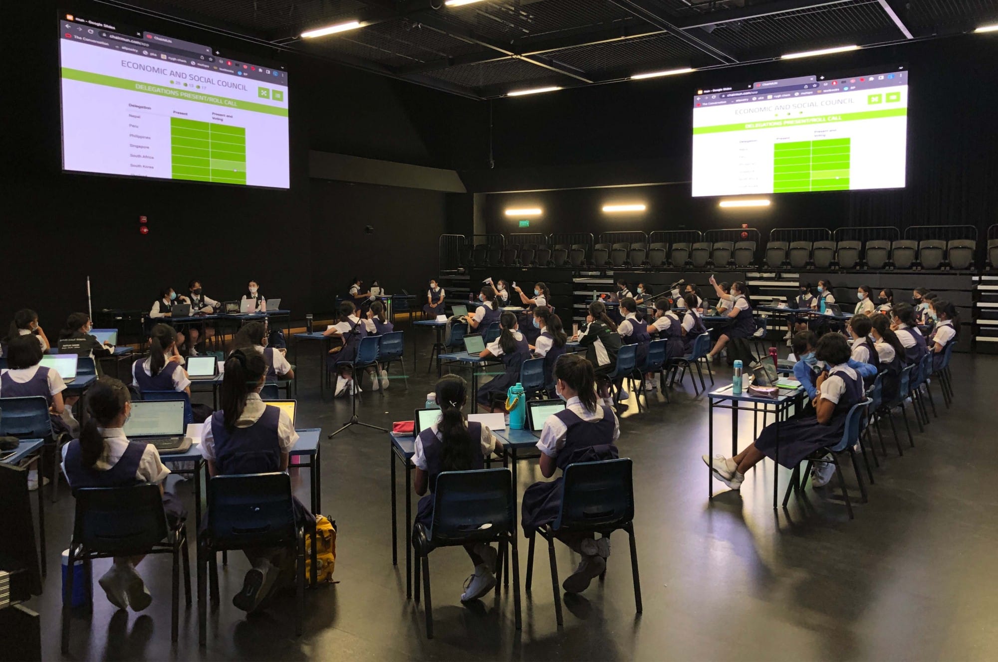 Students in school uniforms sit at desks with laptops in a dark hall, facing two screens showing an Economic and Social Council slide.