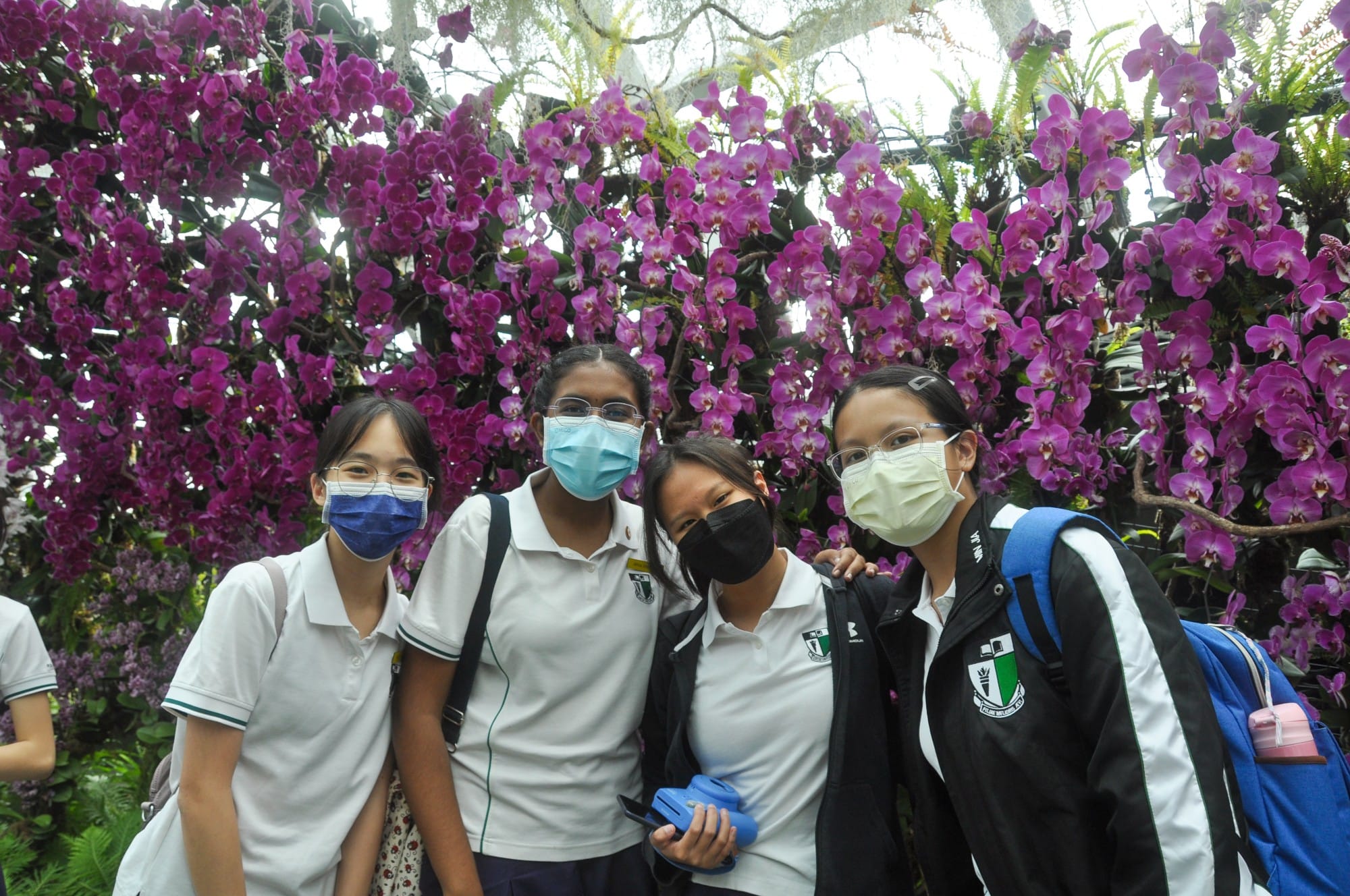 Four masked students in white uniforms pose together in front of a large display of purple orchid flowers.