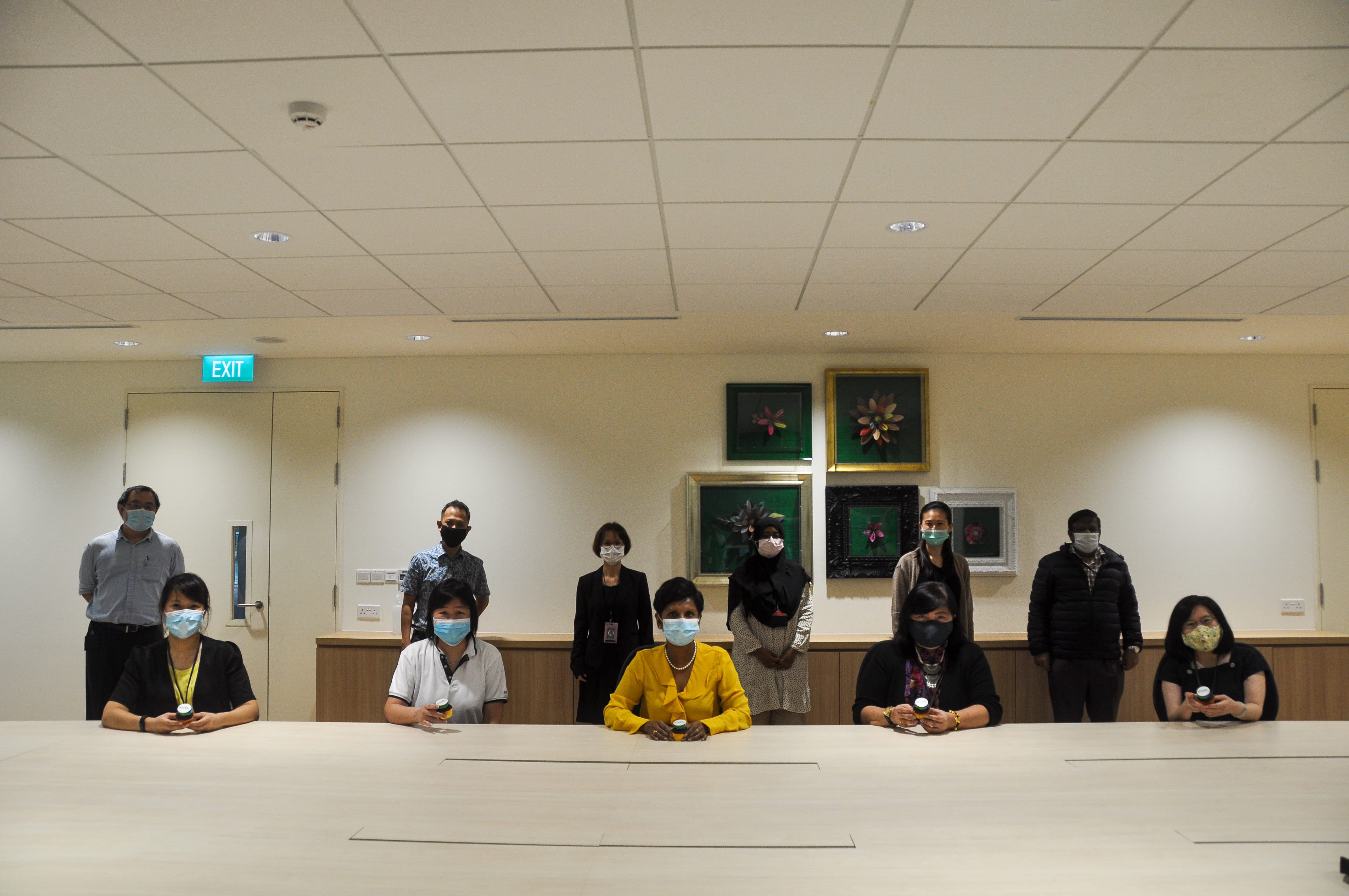 Nine masked people posed in a meeting room behind a long table, with framed flower art on the wall.