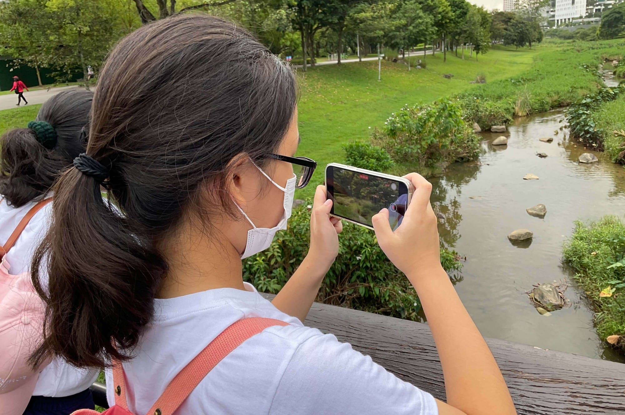 A masked girl with glasses photographs a rocky stream in a green park using a smartphone held horizontally.