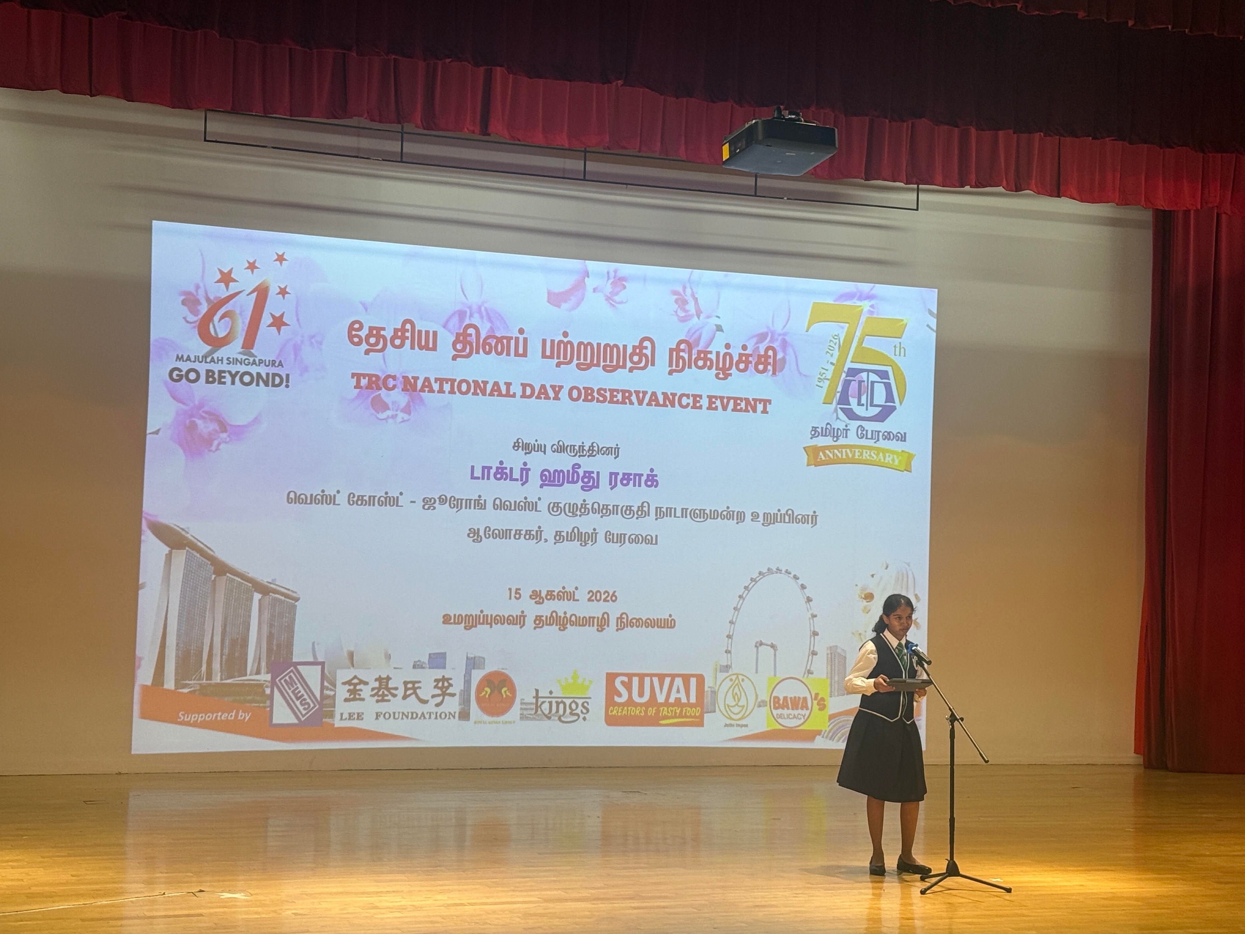 A female student standing on stage in front of a microphone, giving a speech, against a backdrop of a projector screen titled "TRC National Day Observance Event".