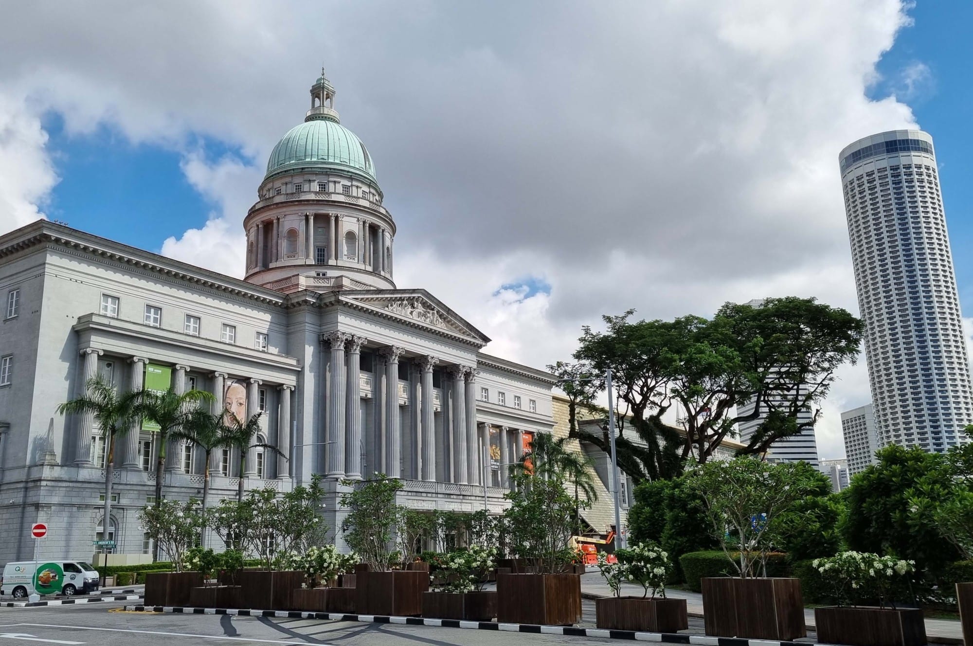 A neoclassical building with a green dome, tall columns, and planters with trees in front, beside a modern skyscraper.
