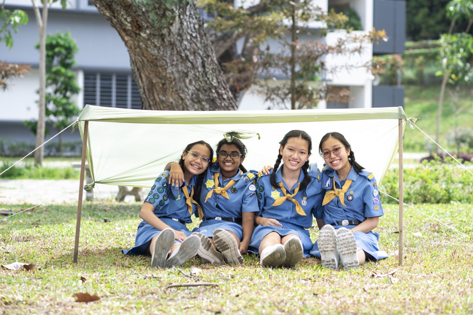 Four smiling girls in blue girl guides uniforms sit on grass under a small green tarp shelter outdoors.