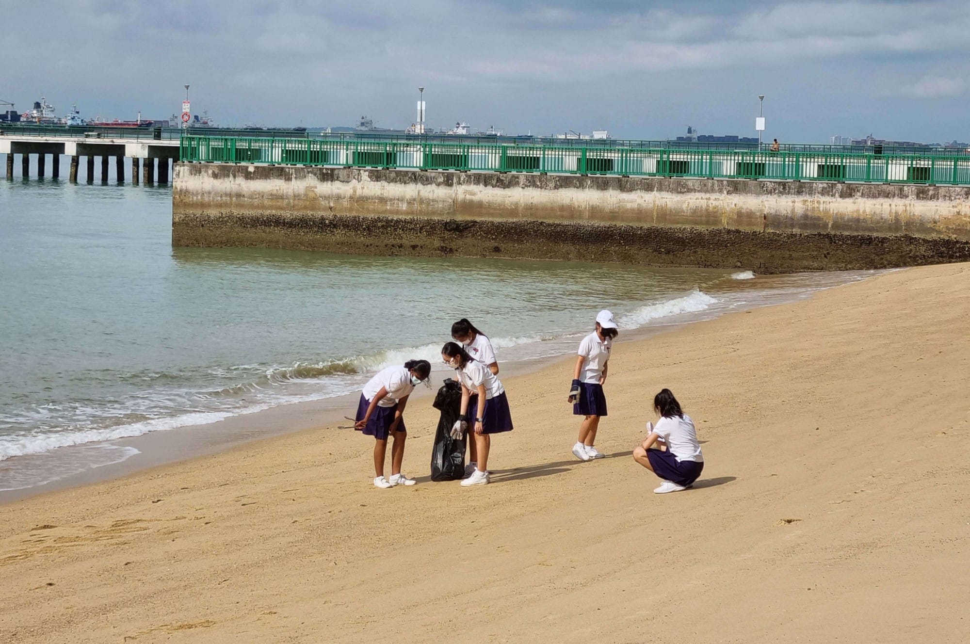 Four girls in white shirts and blue skirts collect trash in a black bag on a sandy beach near a pier.