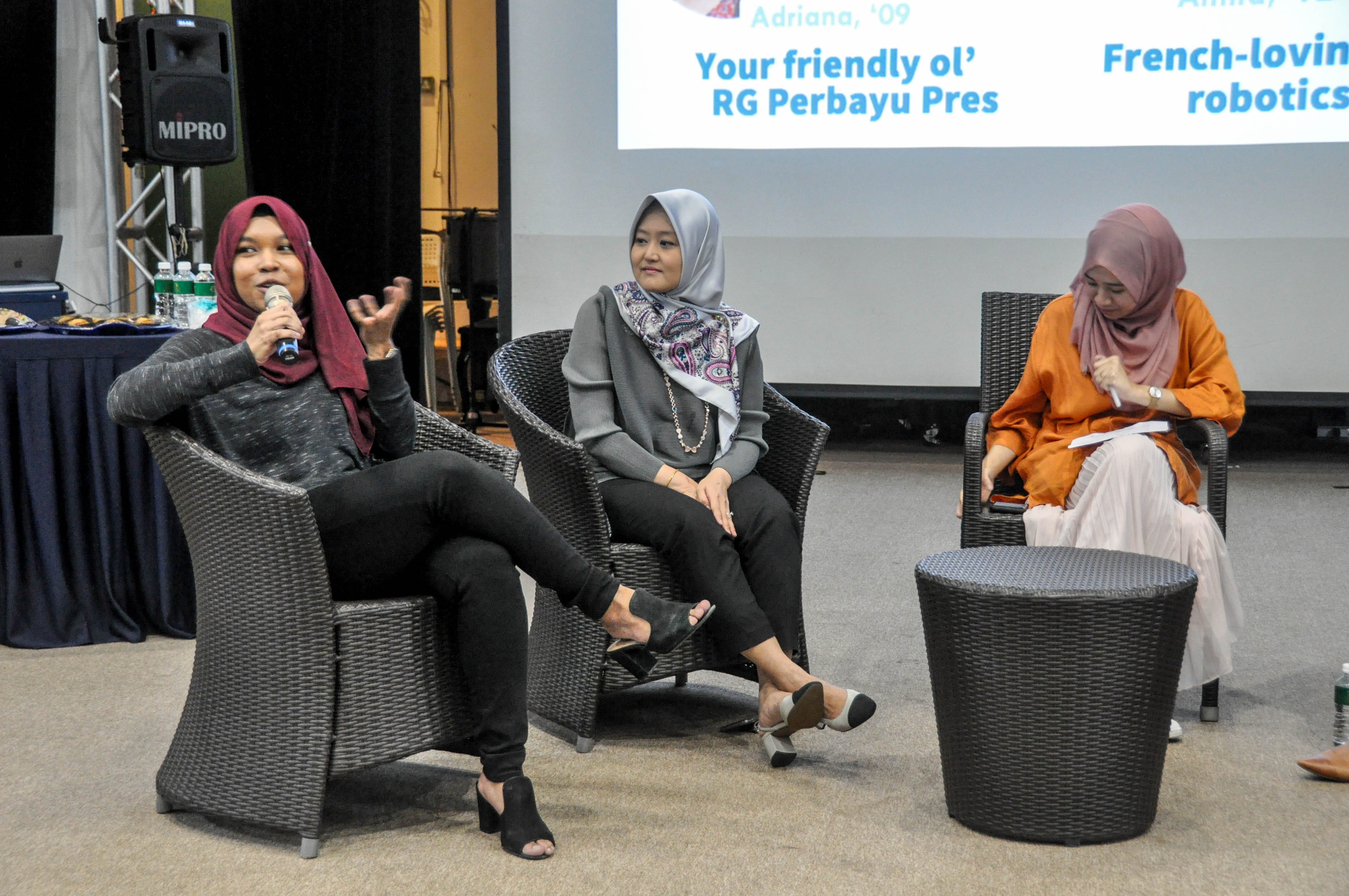Three women wearing hijabs sit in wicker chairs during a panel discussion, one speaking into a microphone.
