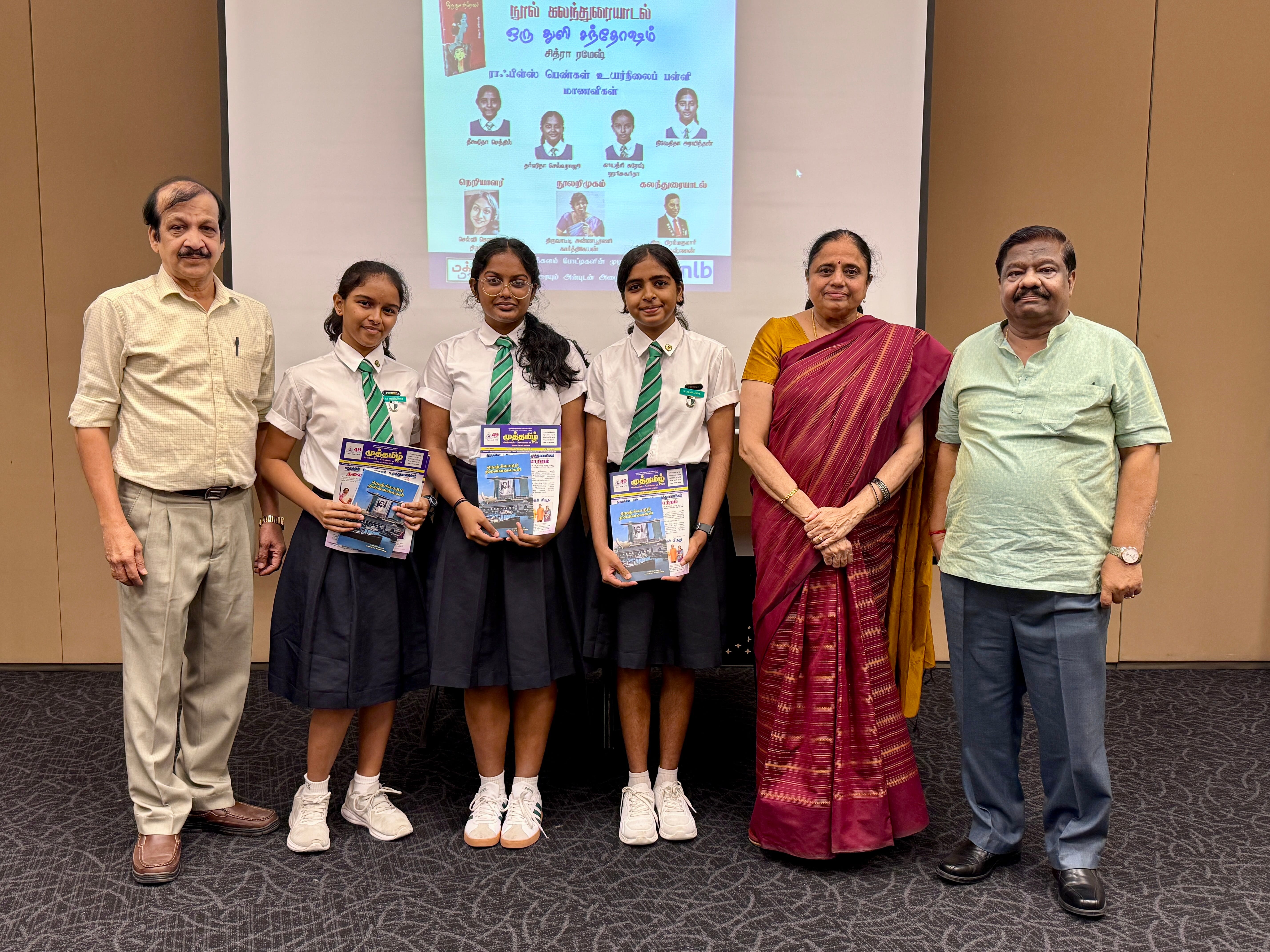 Three schoolgirls in uniform hold books, posing with three adults in front of a projected poster.