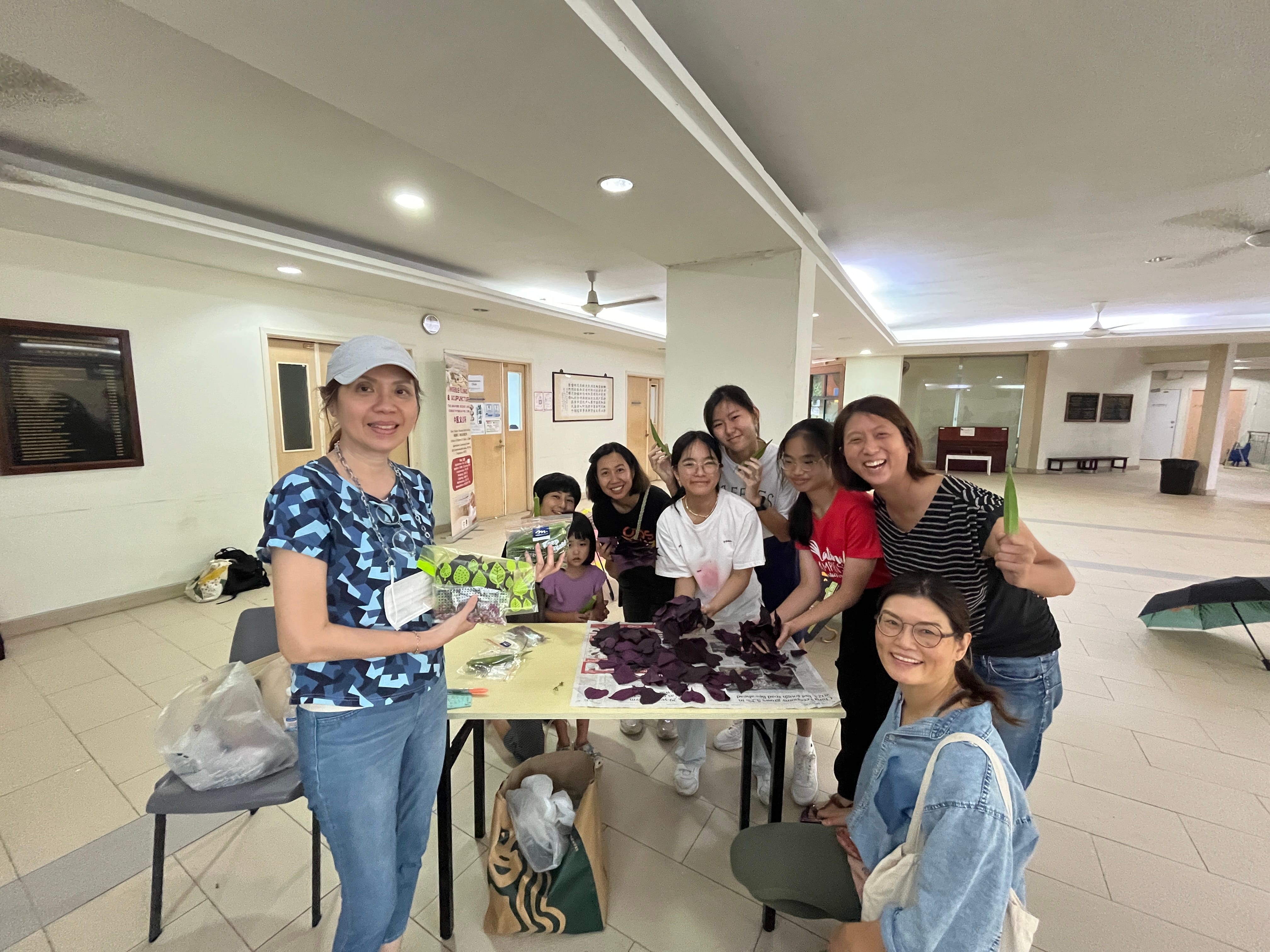 A group of smiling women and children stand around a table covered with purple leaves in an indoor hall.