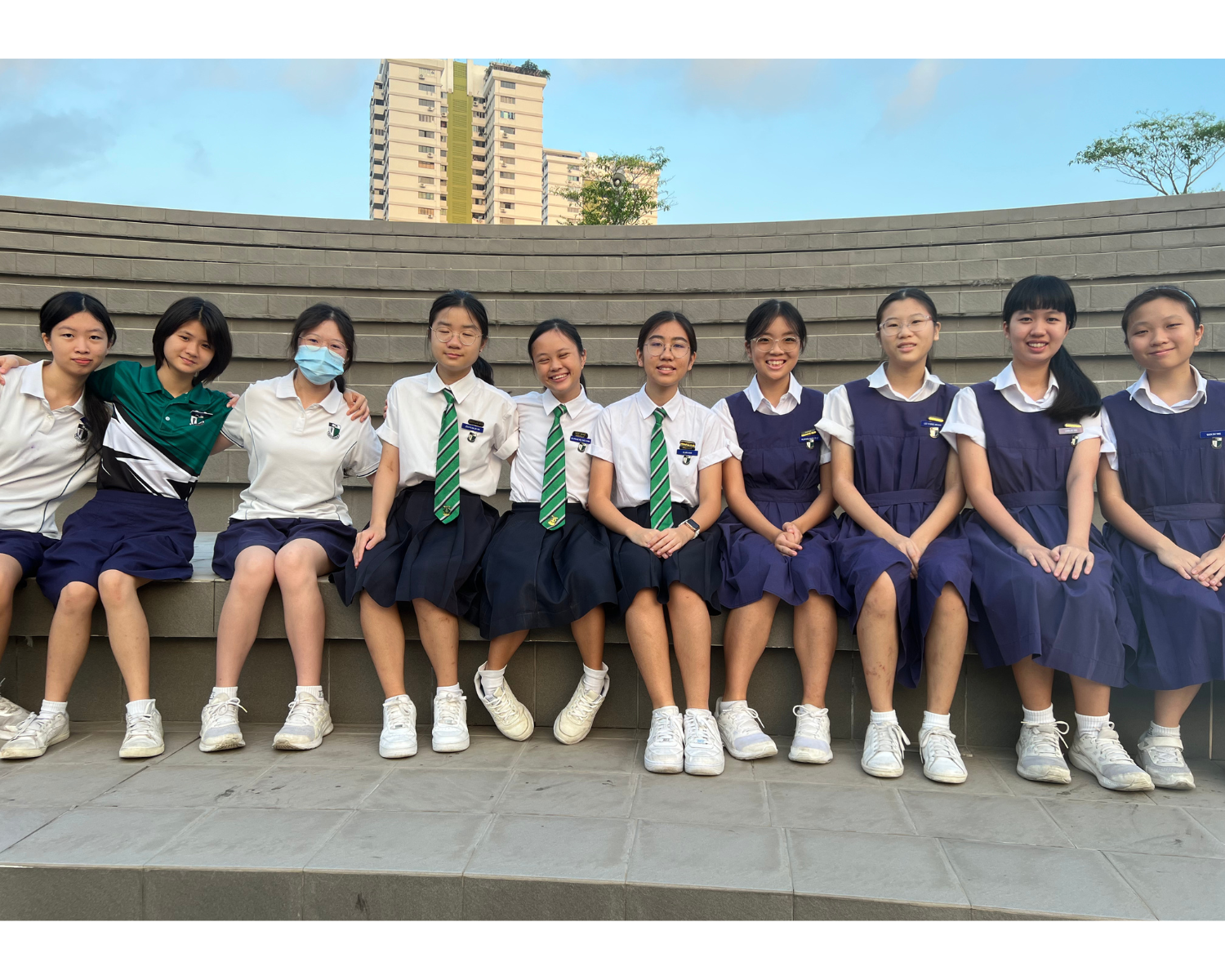 Ten young female students in school uniforms sit together on a curved stone bench outdoors with buildings behind them.