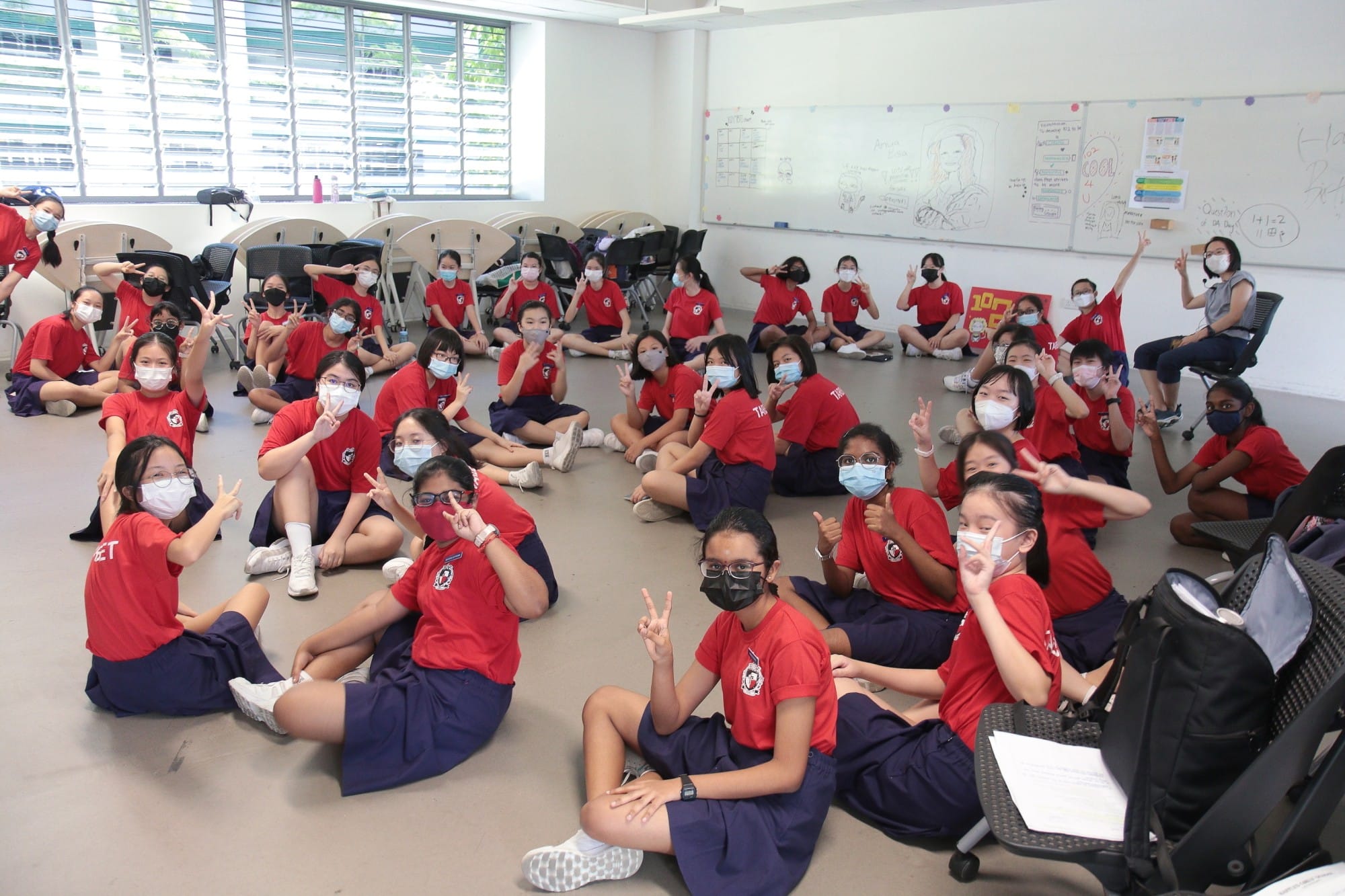 A large group of students in red uniforms and face masks sit on a classroom floor making peace signs.