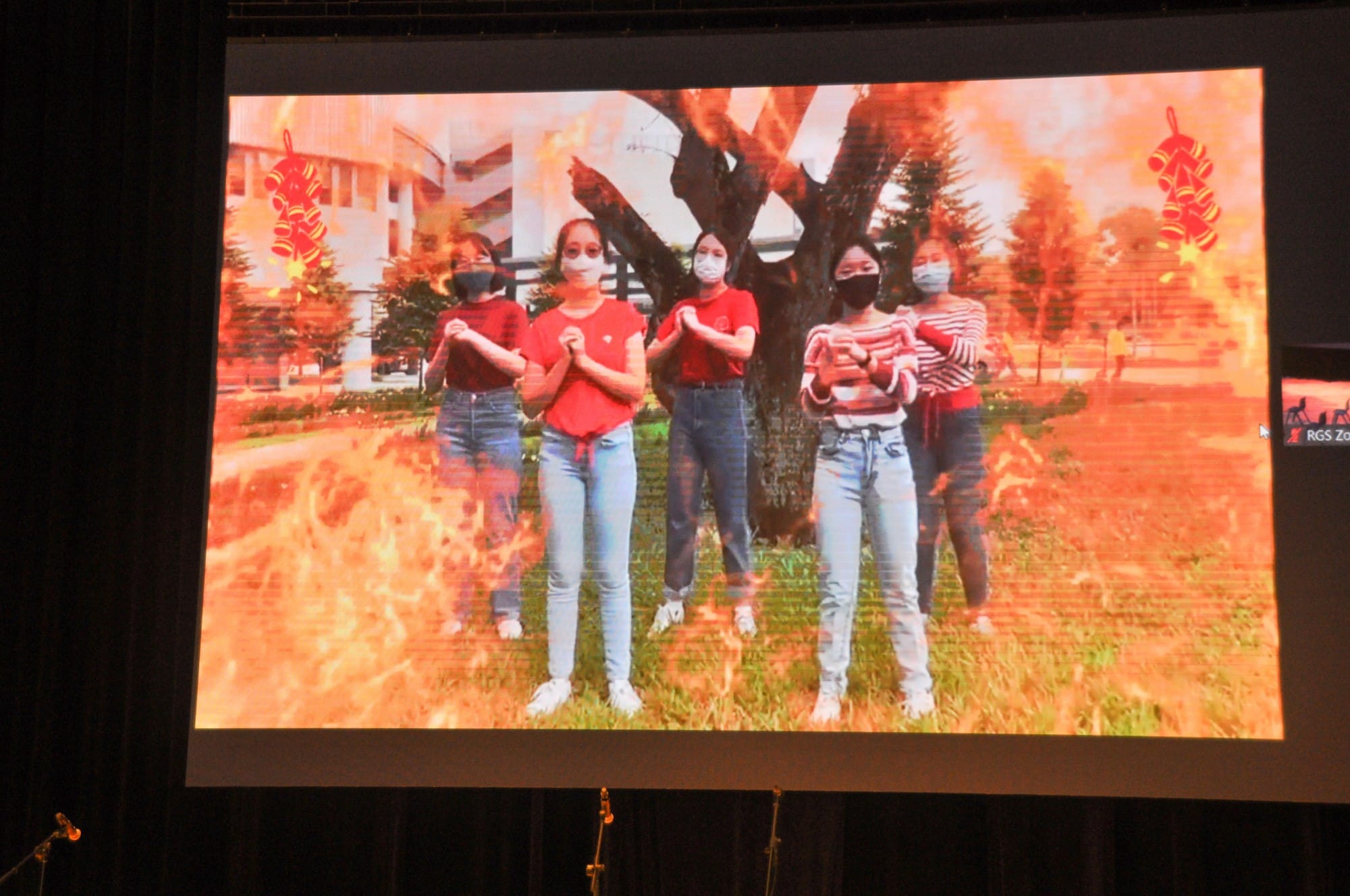Five masked girls in red tops and jeans clasp hands in a Chinese New Year greeting on a projected screen.