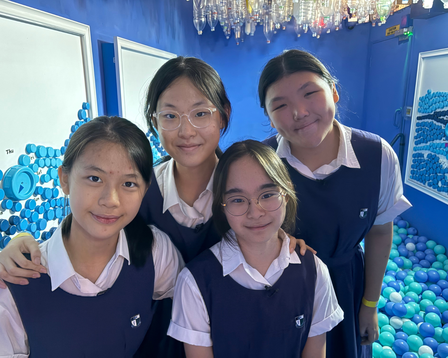 Four smiling schoolgirls in navy uniforms pose together in a blue room with bottle caps and a ball pit.