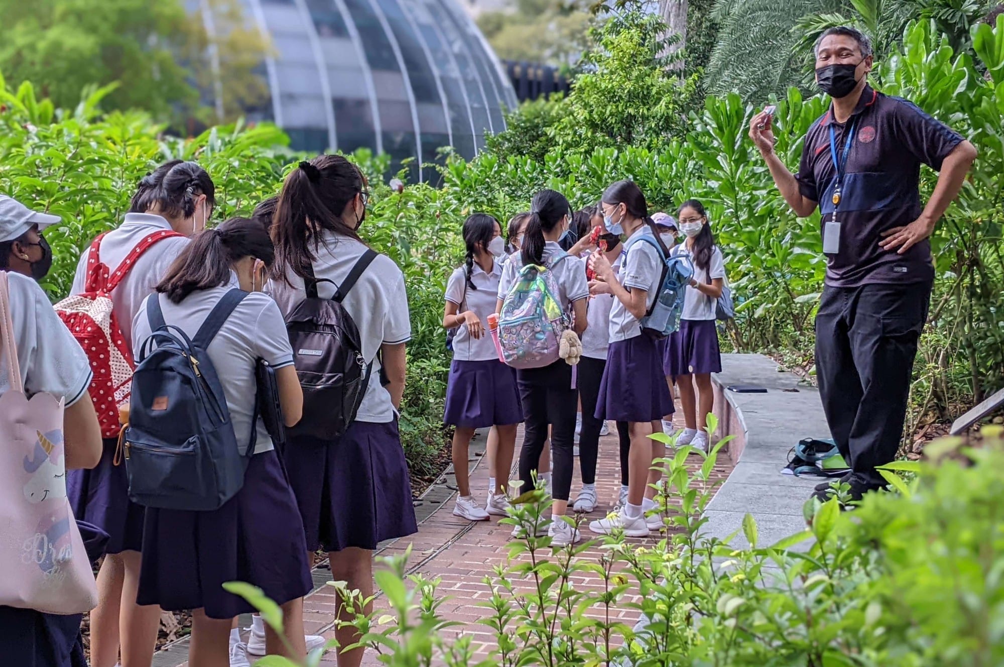 A masked guide speaks to a group of uniformed schoolgirls with backpacks on a garden path near a glass dome.