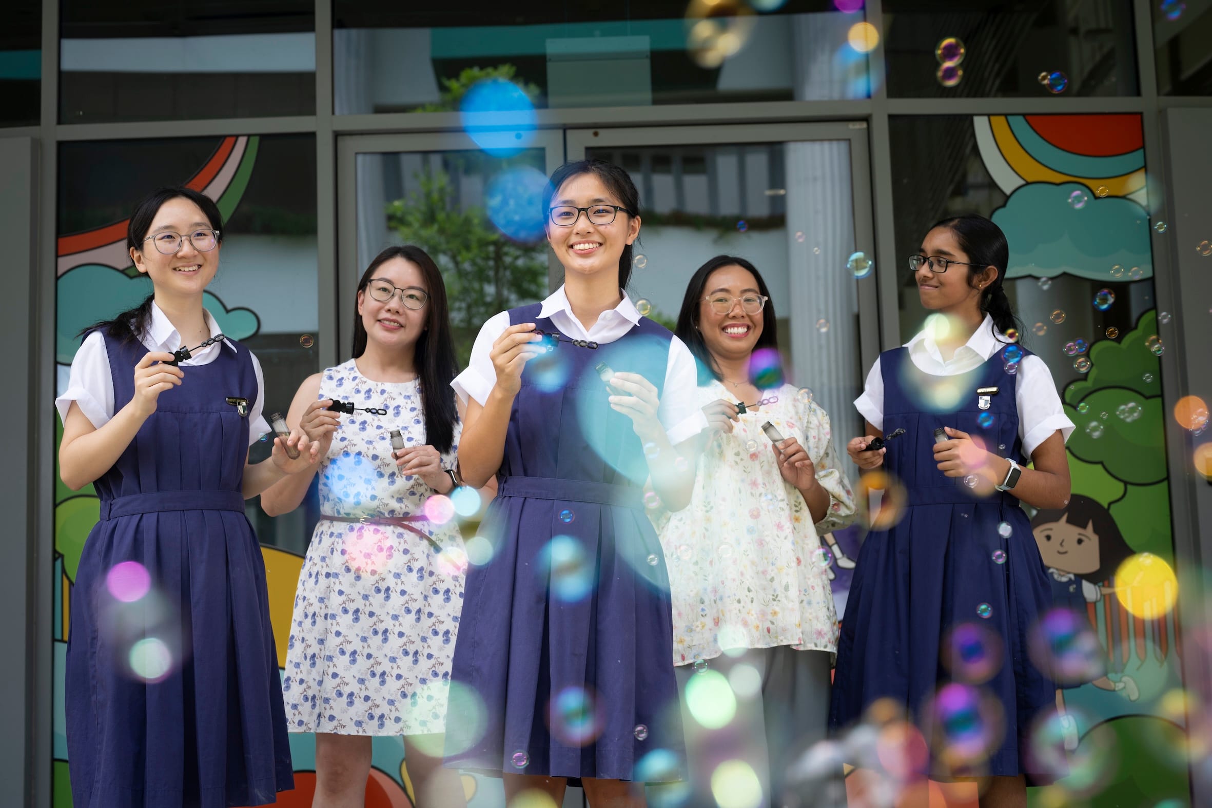 3 students in navy and white uniforms and two other ladies standing in an alternate fashion, blowing bubbles and smiling.