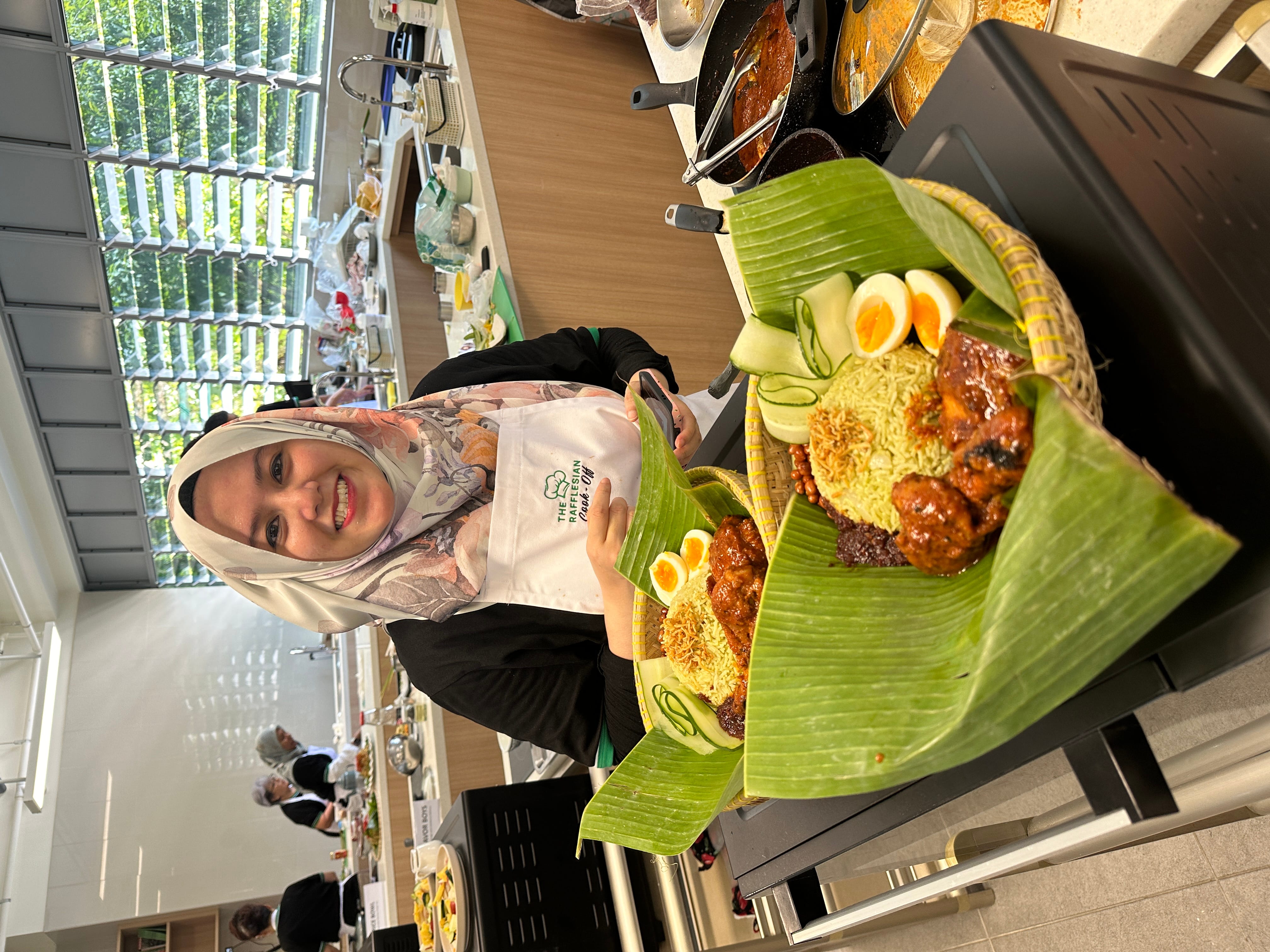 Woman in hijab and white apron smiling, holding baskets of nasi lemak with rice, eggs, and chicken on banana leaves.