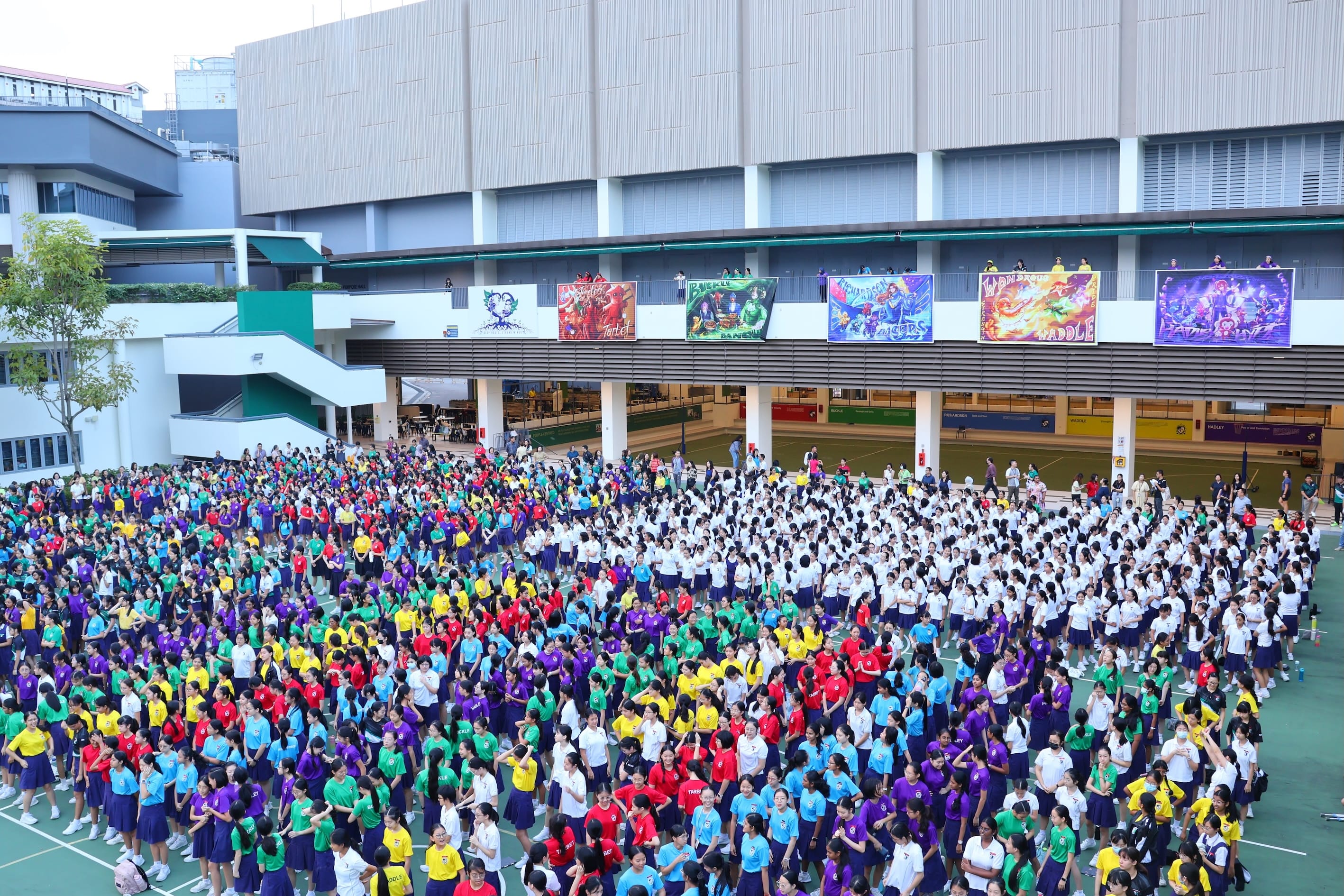 Large crowd of school students in colorful uniforms gathered in an outdoor courtyard with colorful banners overhead.
