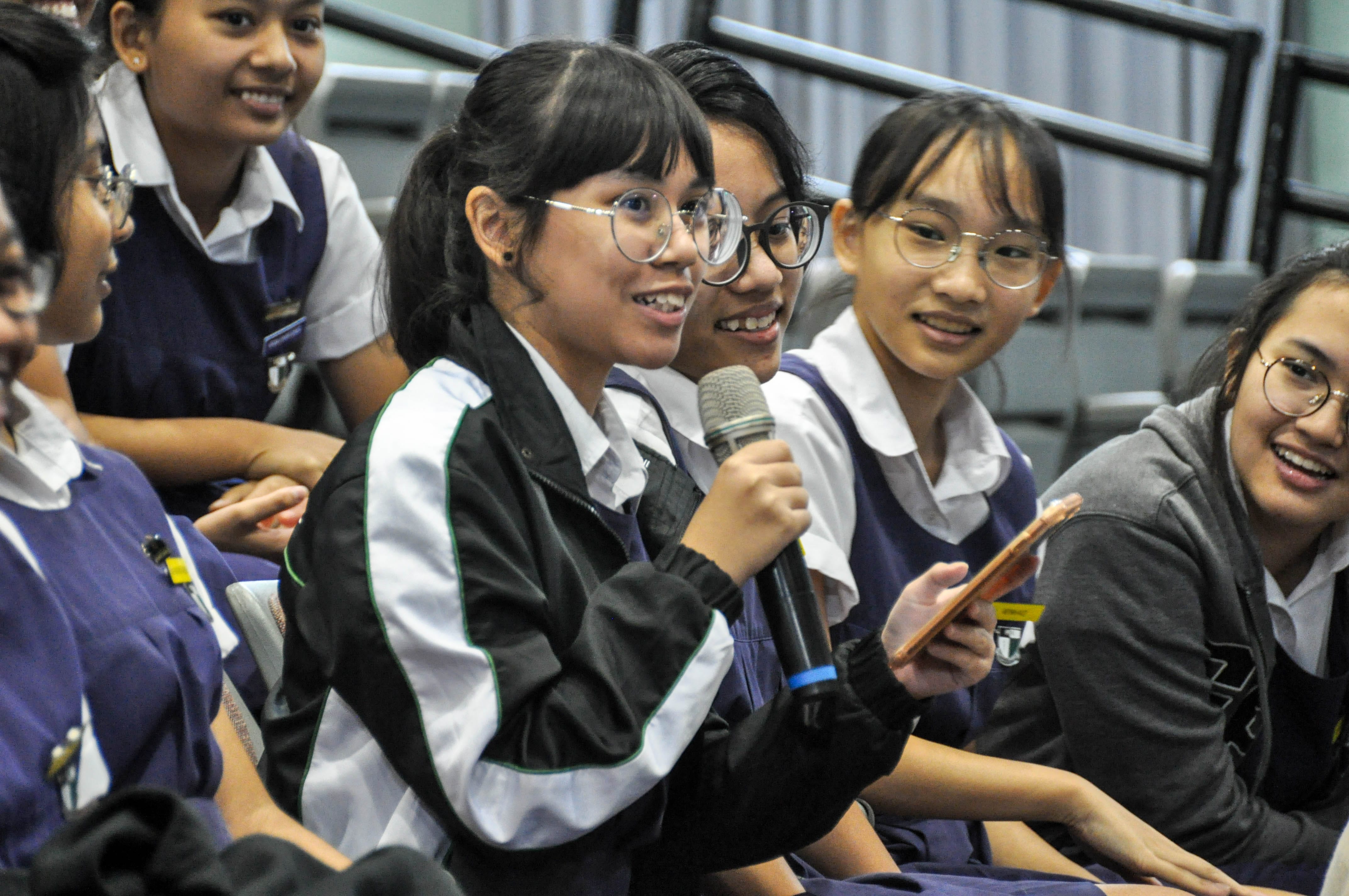 A smiling teenage girl in glasses holds a microphone while seated among other uniformed students.