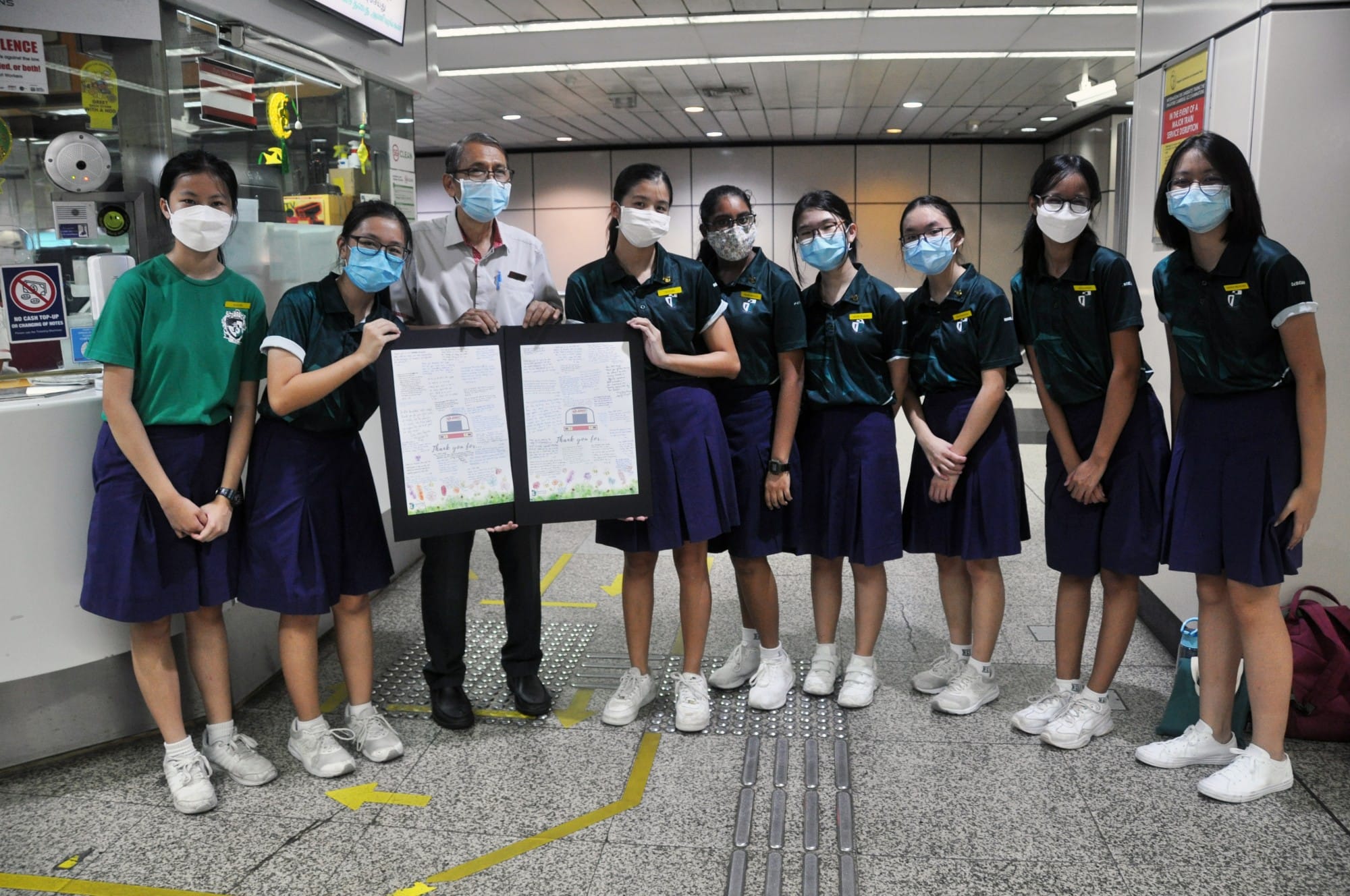 A group of masked schoolgirls and an adult man pose together holding a framed thank-you poster inside a transit station.