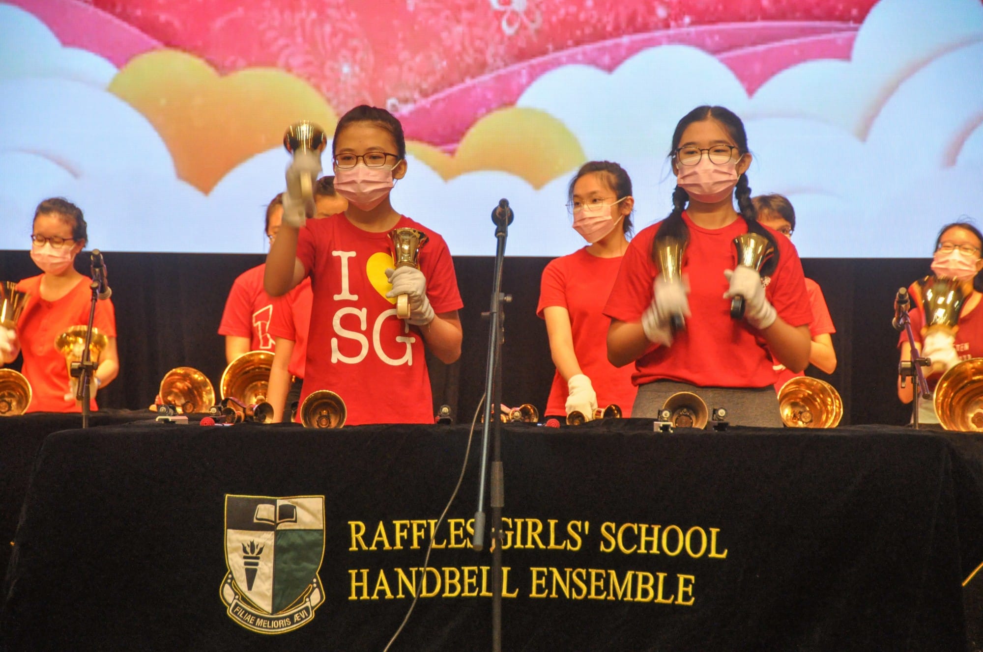 Girls in red shirts and masks perform with handbells on stage for Raffles Girls' School Handbell Ensemble.