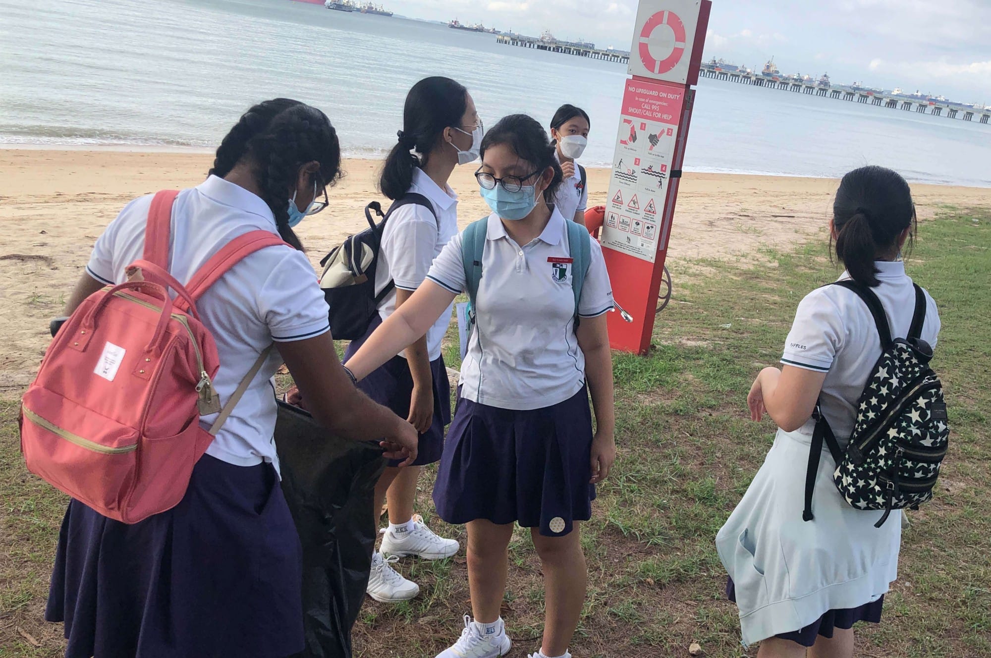 Several masked schoolgirls in uniforms with backpacks stand near a beach safety sign beside a sandy shore.