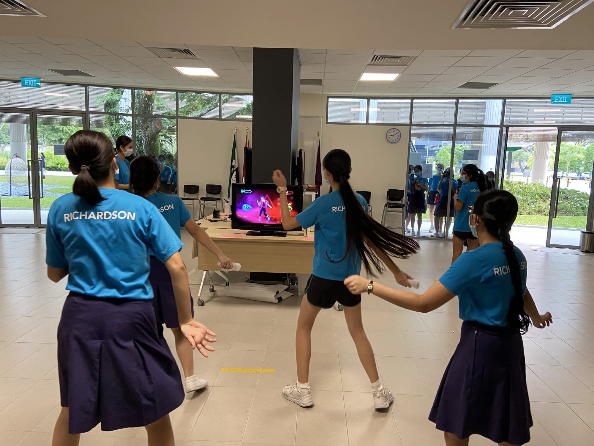Students in blue Richardson shirts and navy skirts play Just Dance on a TV screen inside a bright school lobby.