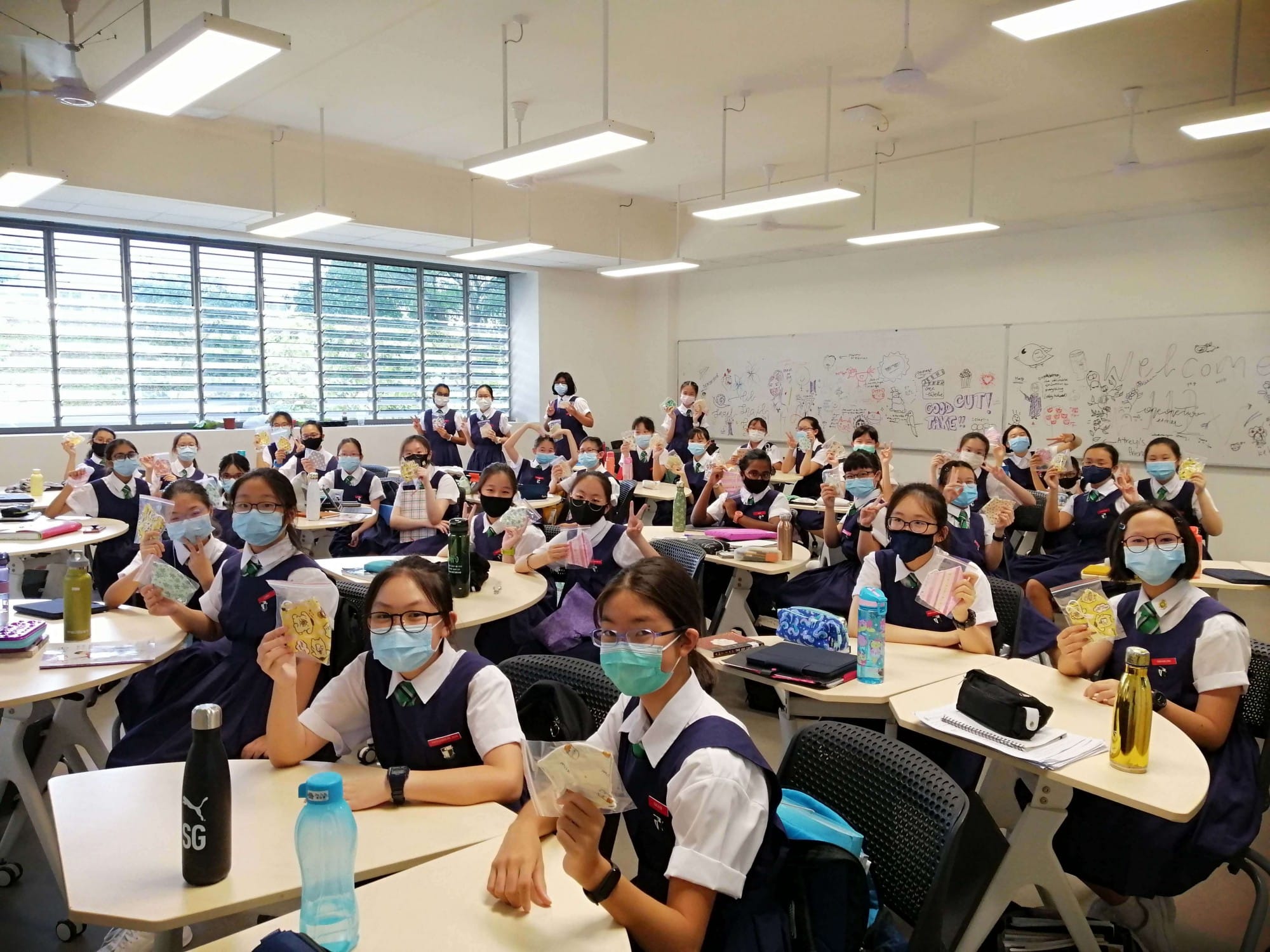 Students in navy uniforms and face masks sit at desks in a bright classroom, holding up small cards or crafts.