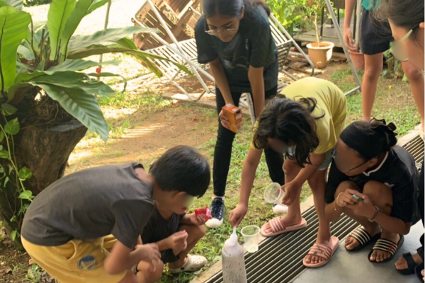 Several children and teens gather outdoors around a bottle, pouring liquids in what appears to be an experiment.