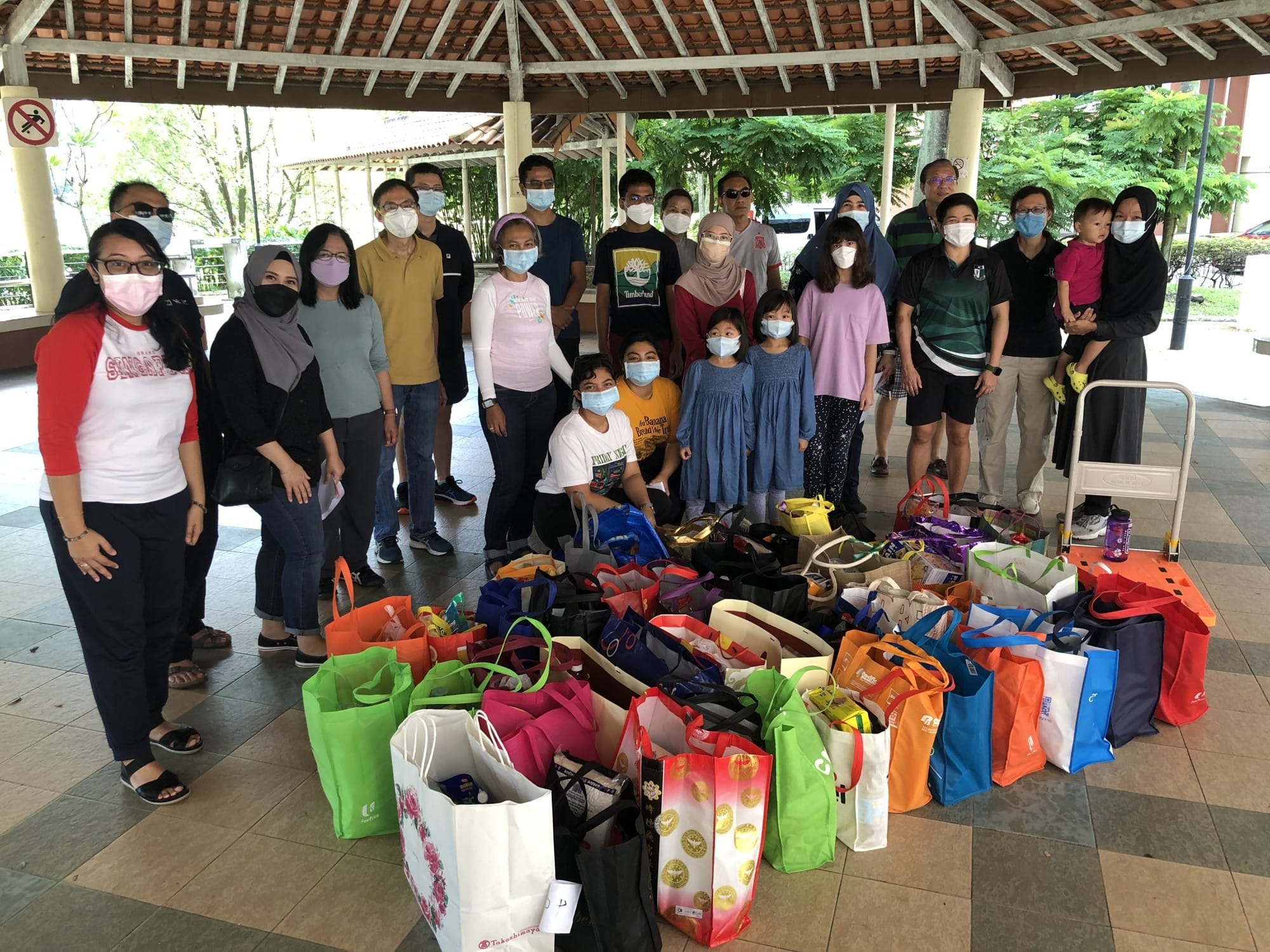 A large group of masked adults and children stand under an outdoor pavilion surrounded by many colorful donation bags.