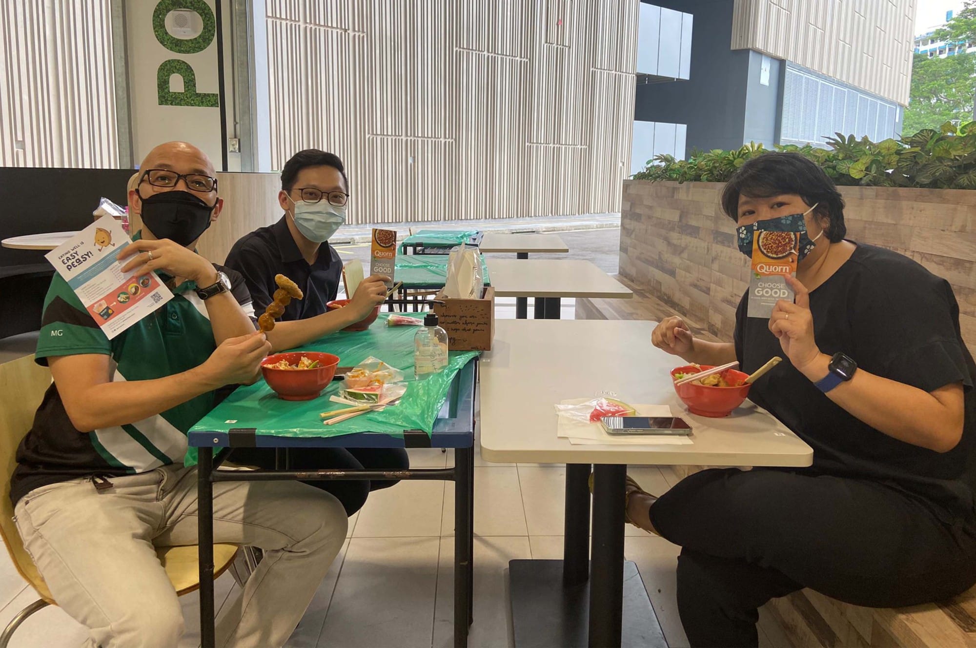 Three masked people sit at tables with red bowls of food, holding up Quorn product boxes and flyers.
