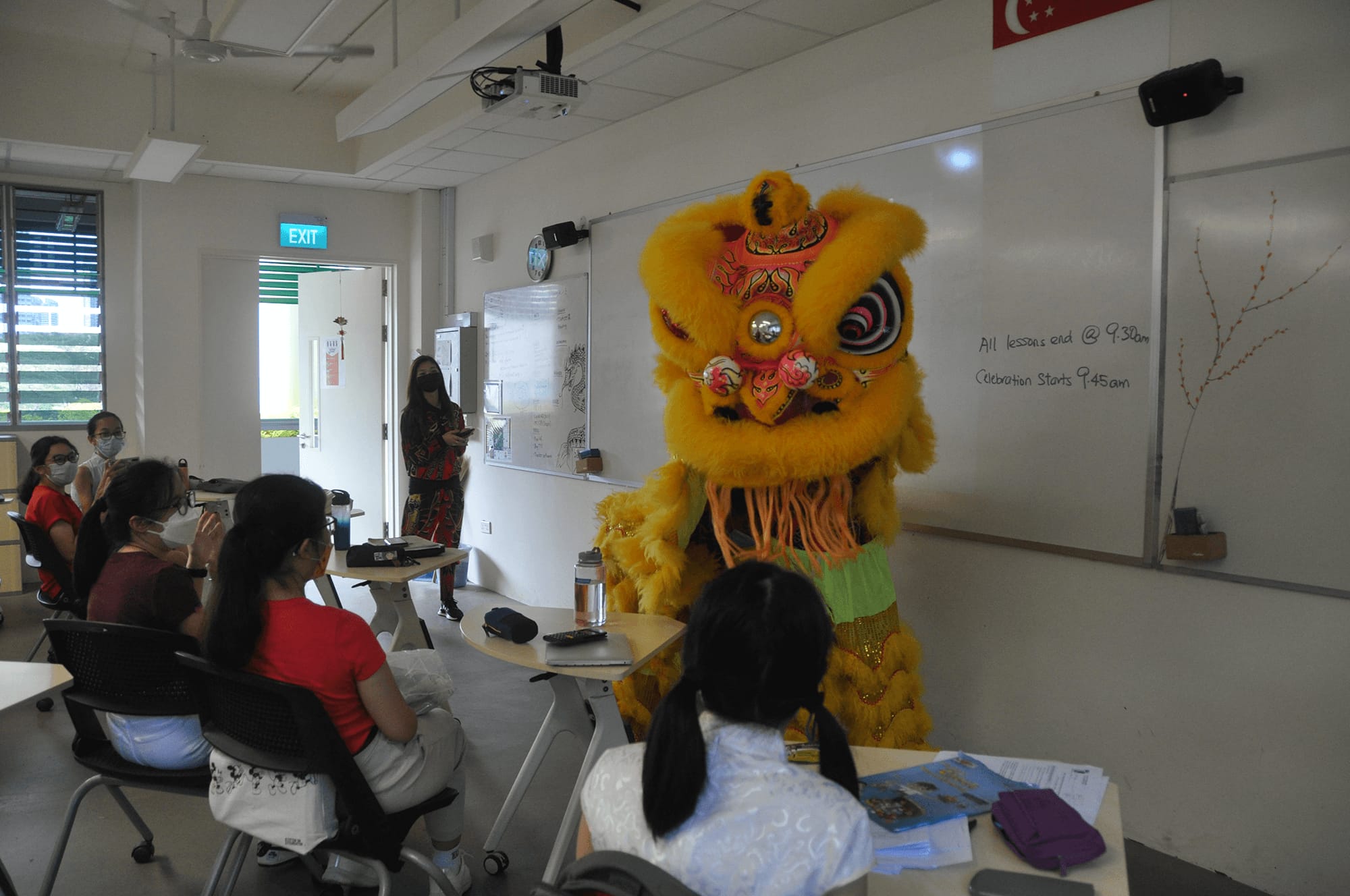 A bright yellow Chinese lion dance costume performs in a classroom while students seated at desks watch.
