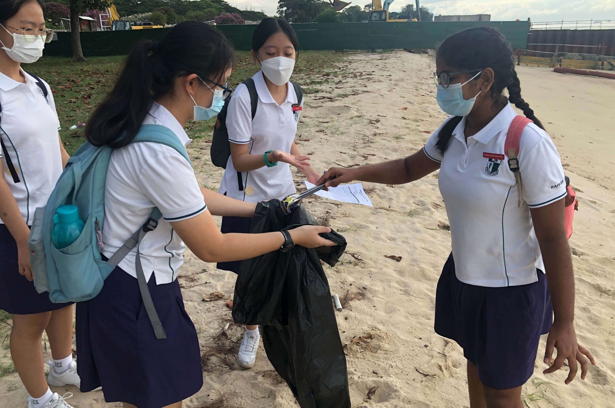 Four masked students in white uniforms collect trash in a black bag on a sandy beach.