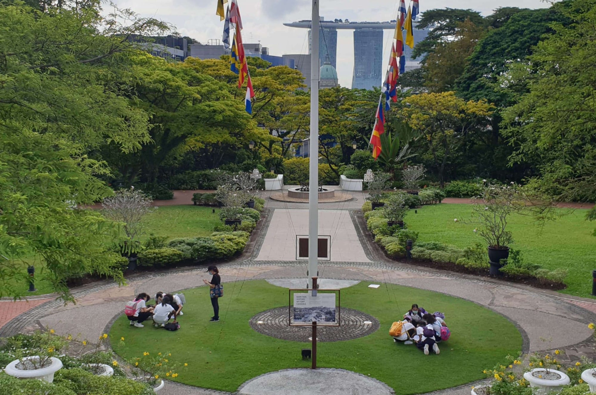 A formal garden with a central flagpole, colorful flags, green lawns, and groups of students sitting on the grass.