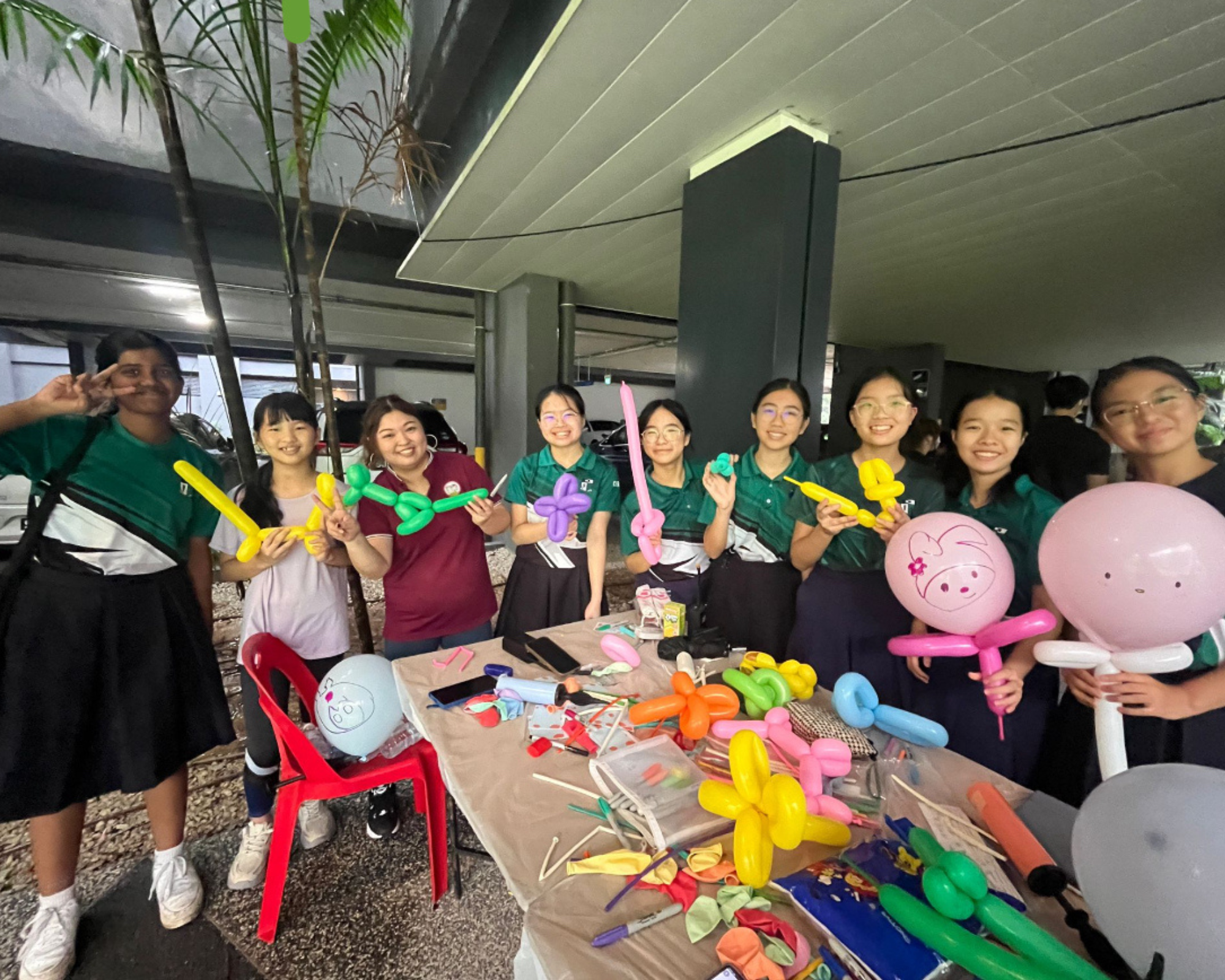 Group of smiling students by a table with balloon animals and pink balloon faces in a covered outdoor area
