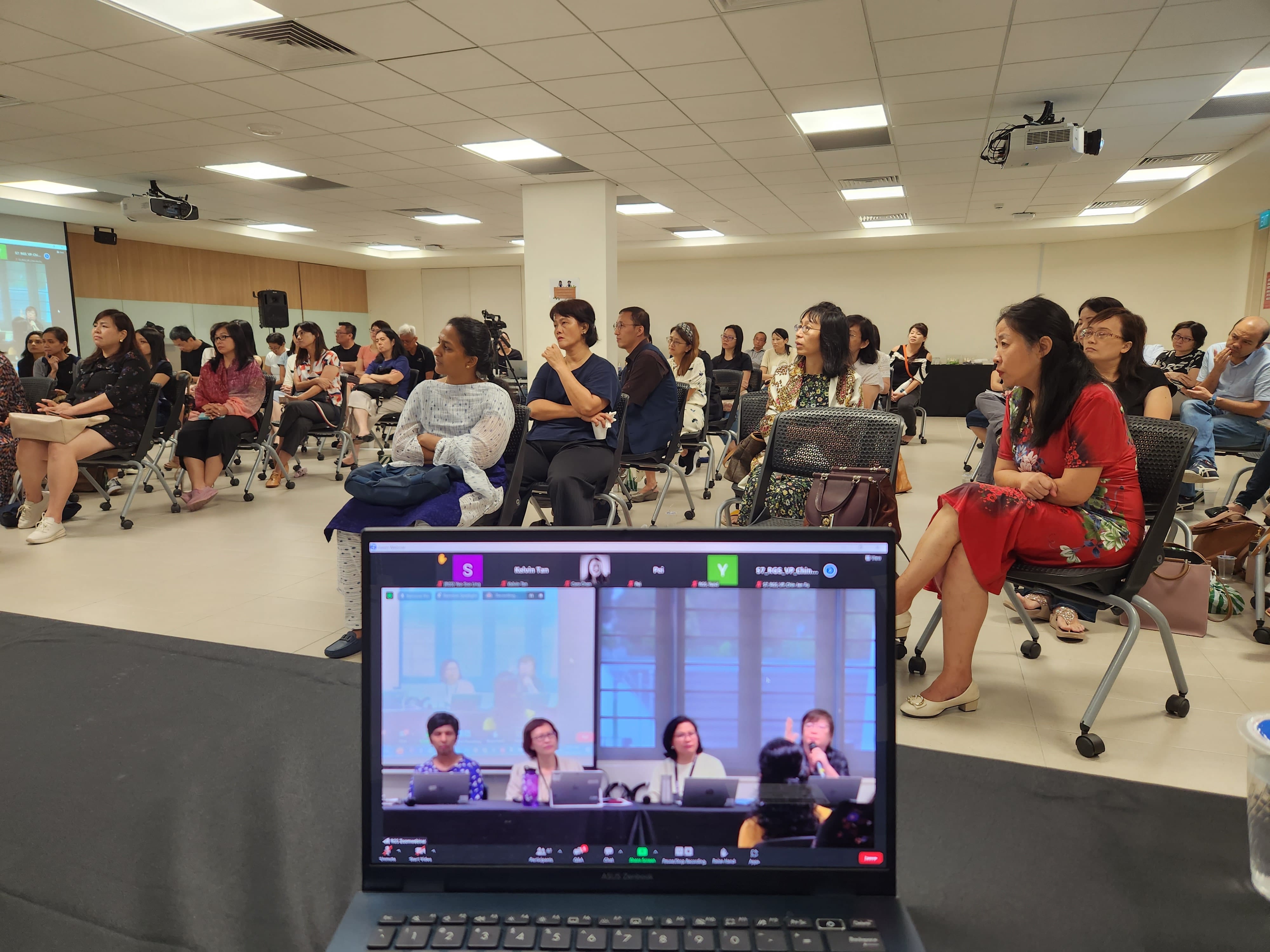 A large group of people seated in a conference room watching a presentation, with a laptop showing a Zoom meeting in the foreground.