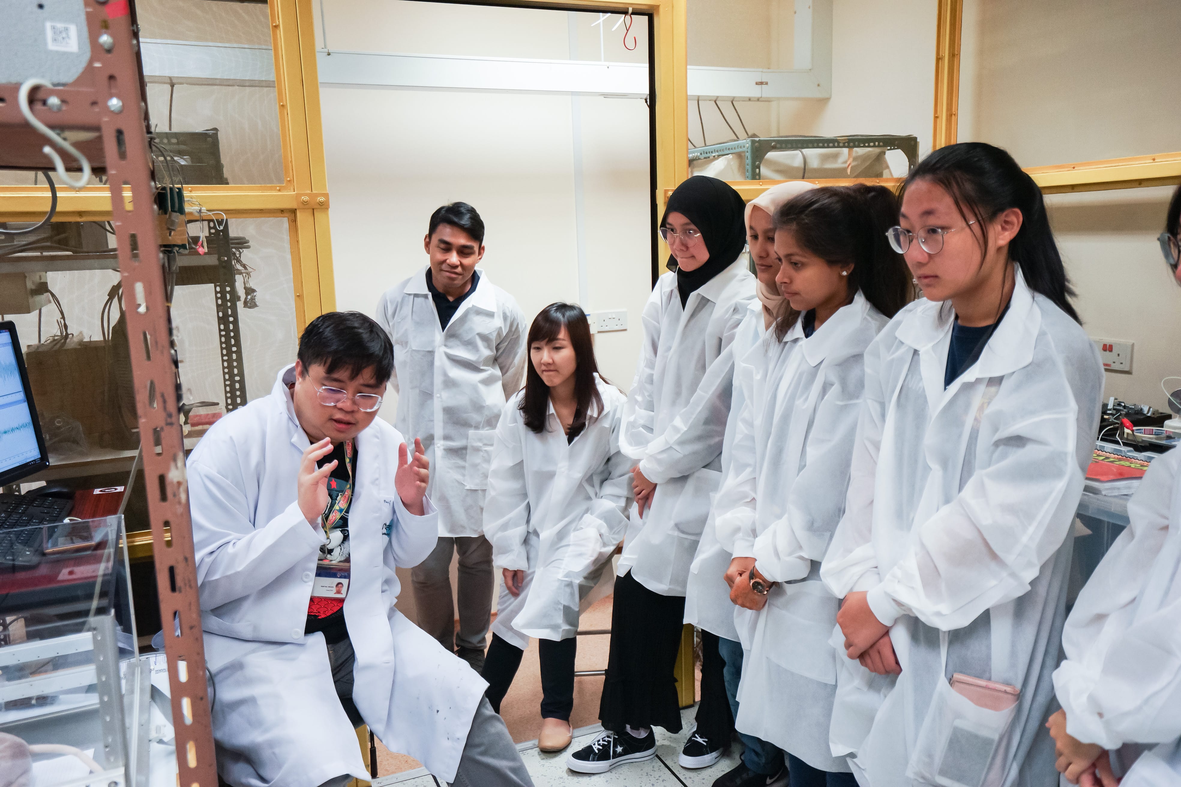 A researcher in a white lab coat gestures while explaining to a group of students in white coats inside a laboratory.